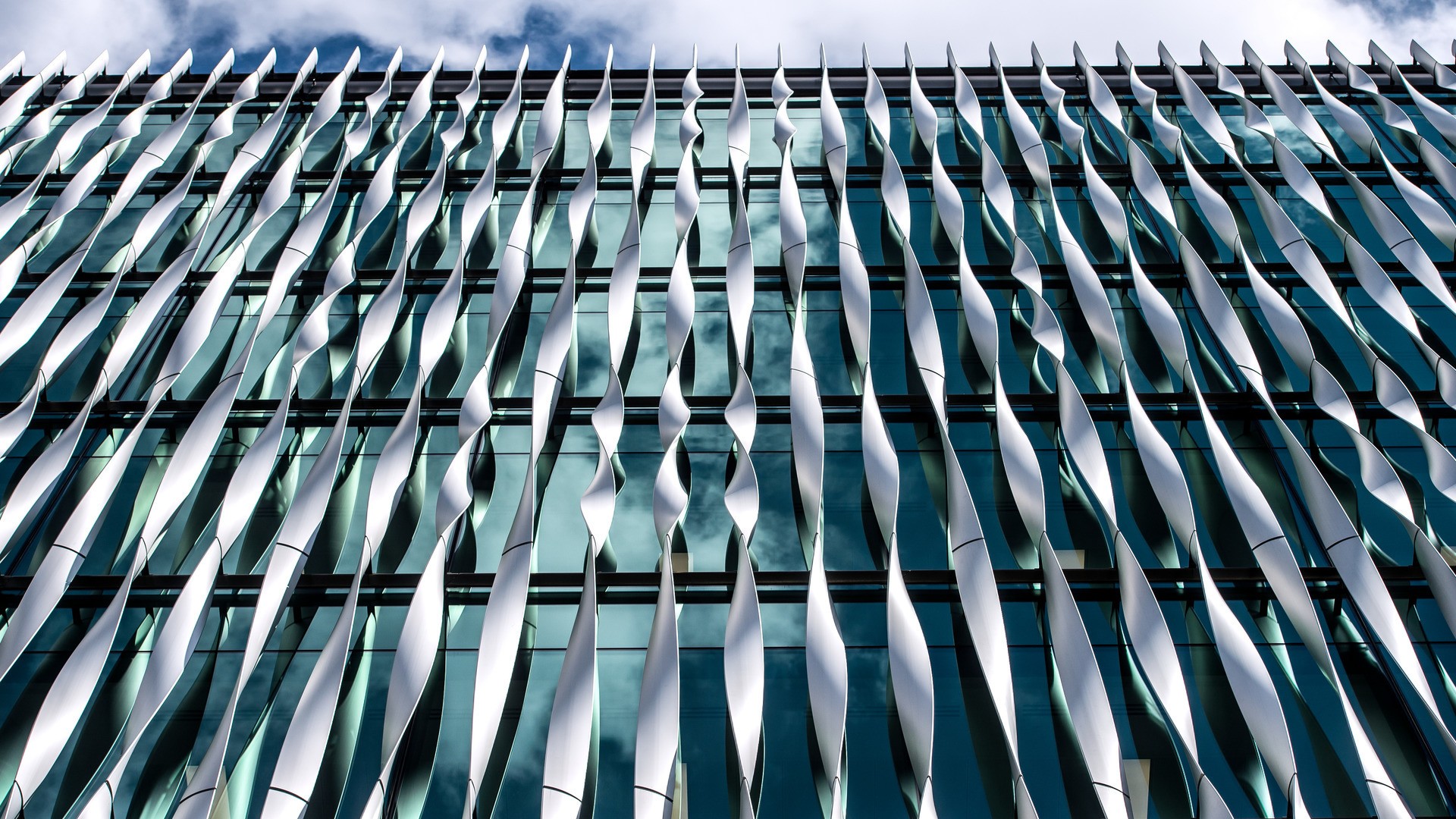 Modern building facade with twisting metal fins and reflective glass windows under a cloudy sky.