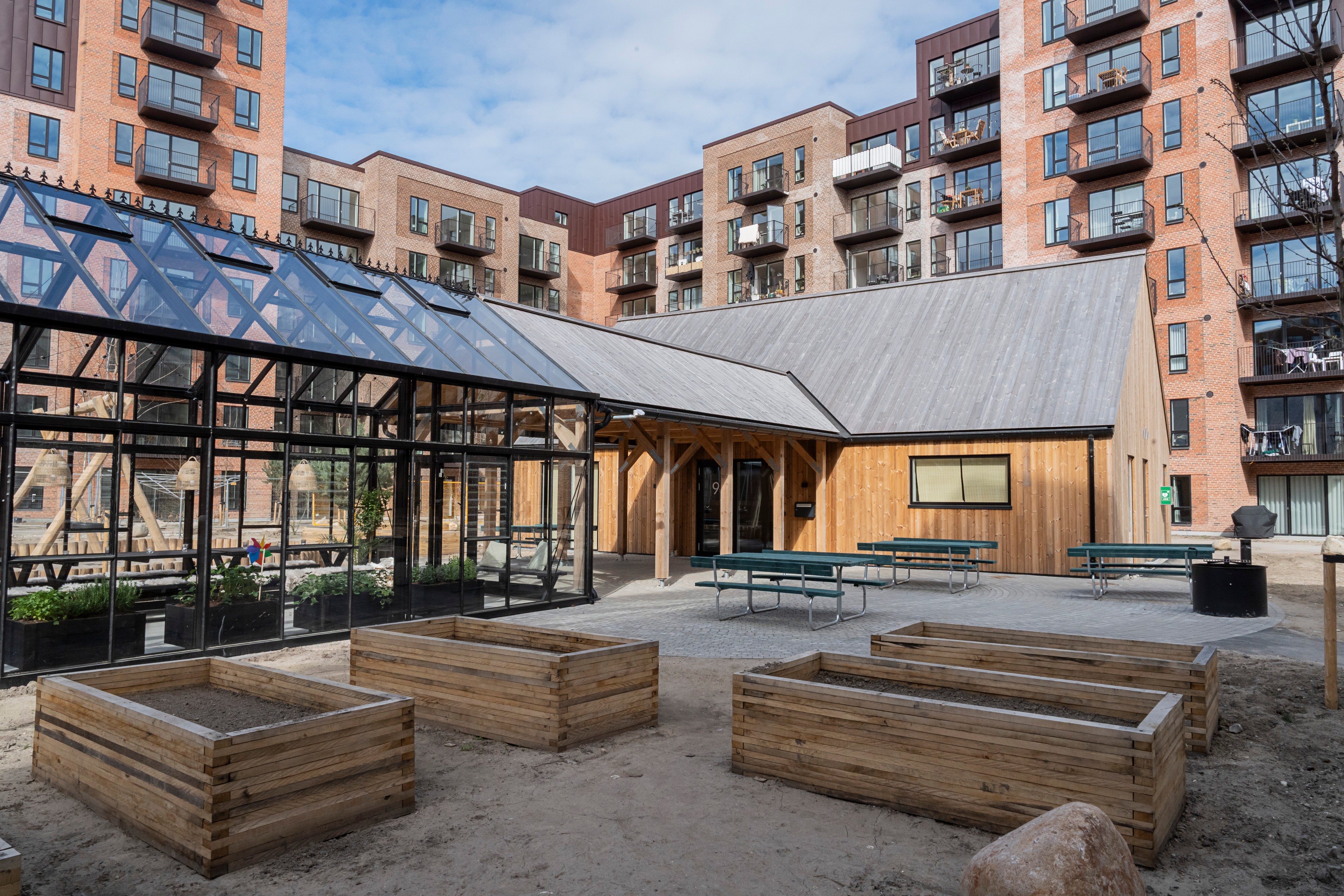 Urban community garden with greenhouse and raised beds surrounded by modern apartment buildings.