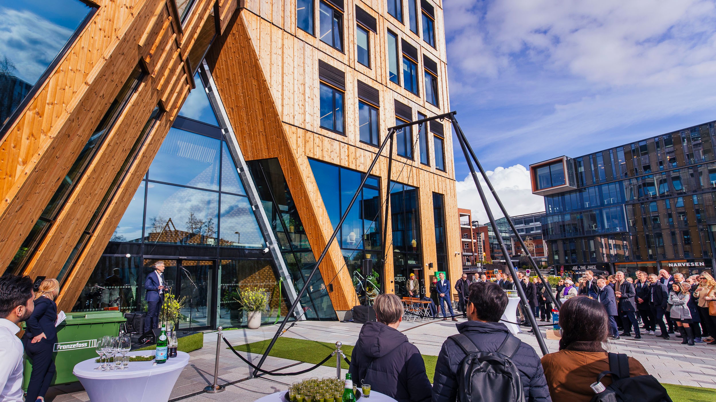 Outdoor event with people gathered near modern wooden and glass building under blue sky.