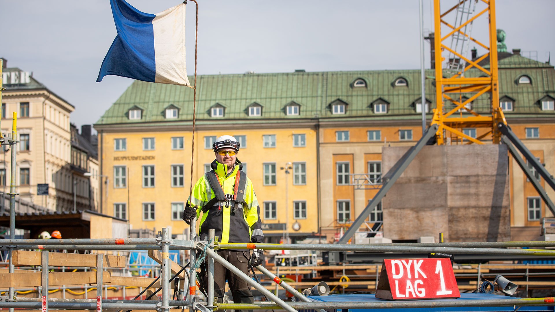 Säkerhetsarbete vid Slussen Martin Johansson står med full säkerhetsmundering vid den hissade blåvita dykarflaggan som visar att det är dykare i vattnet. På pråmen står det dyklag 1.