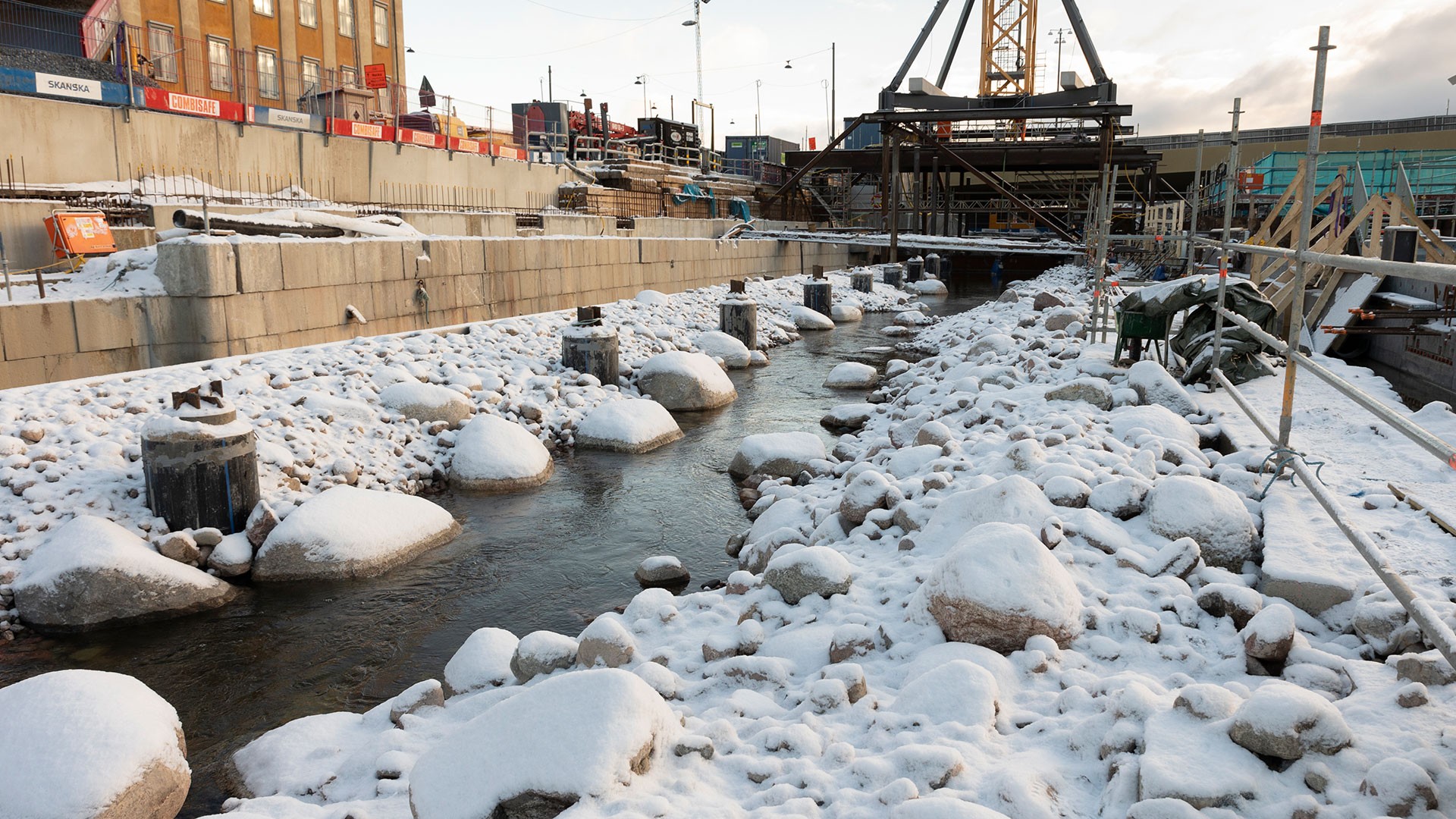 Snötäckt byggarbetsplats med en smal bäck, omgiven av stenar och industrimaskiner.