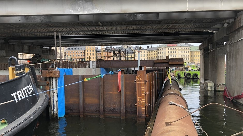 Under en bro, ett kajområde med en båt som heter TRITON, vatten och industristrukturer.