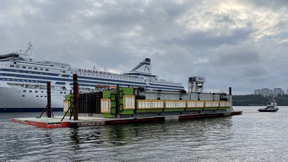 Stort kryssningsfartyg förtöjt nära en flytande plattform med containrar under en molnig himmel.