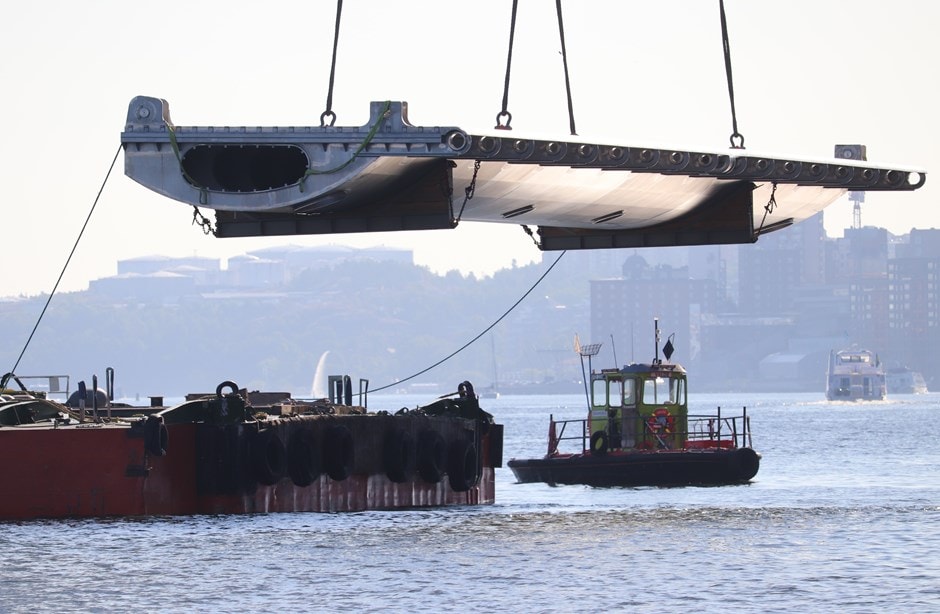 En stor metallbrosektion lyfts av kranar över vatten nära en bogserbåt och pråm.