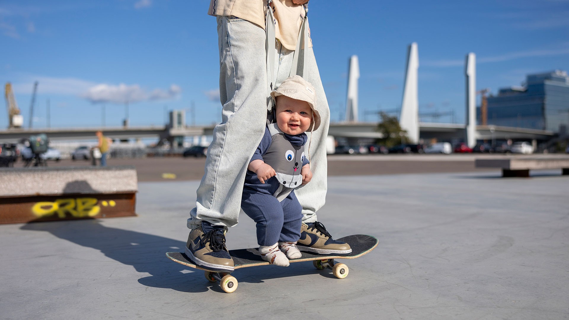 En vuxen och ett litet barn delar en skateboardtur i en stadspark under en klarblå himmel.