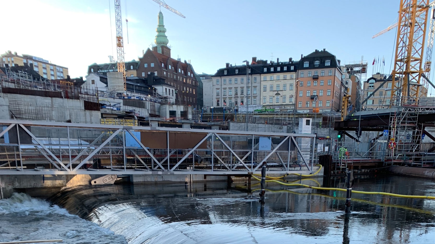 Urban byggarbetsplats med kranar, byggnader och en bro över en flytande vattenväg i staden.