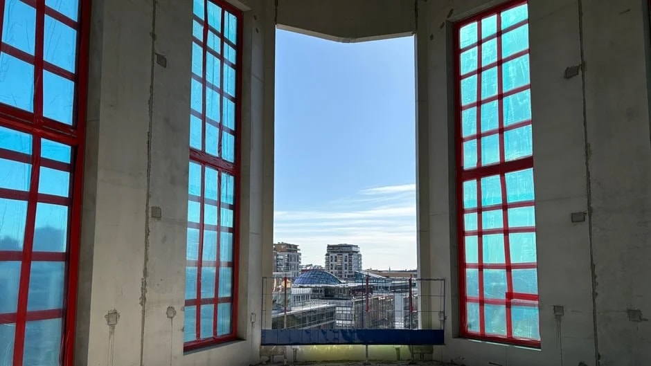 Interiörvy av en byggnad under konstruktion med stora fönster och utsikt över stadssilhuetten.