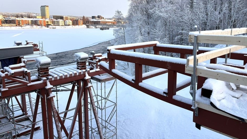Snötäckt stålkonstruktion och byggnadsställning vid en flodbank med snötäckta träd och byggnader.