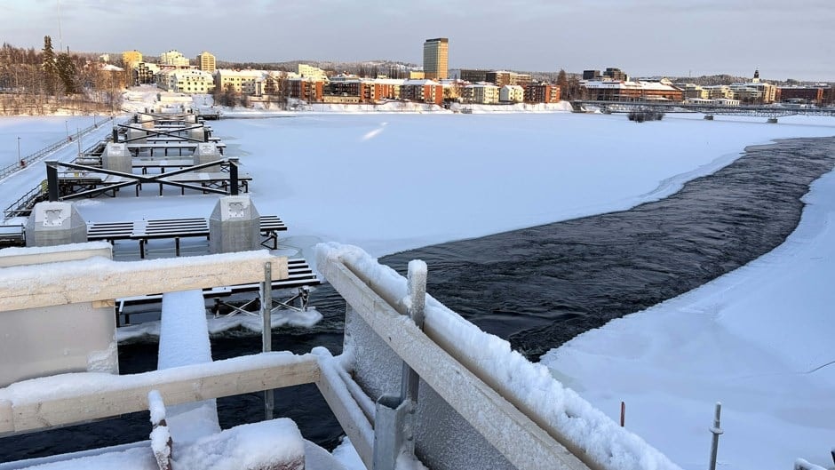 Snötäckt bro över en delvis frusen flod med en stadssilhuett i bakgrunden.