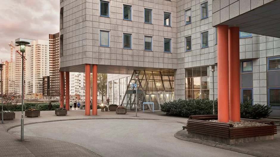 Modern stadbyggnad med stora fönster, orangea pelare och omgivande grönska.