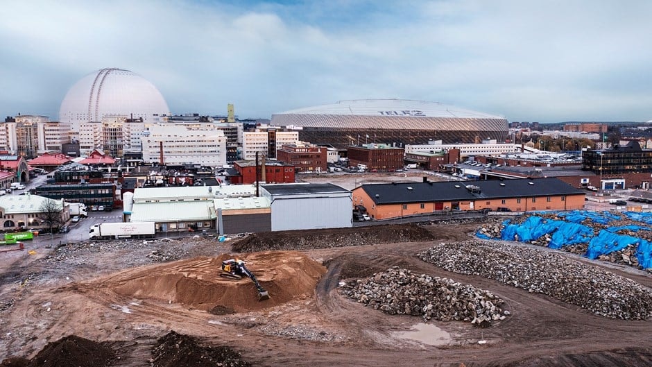 Byggarbetsplats med maskiner i förgrunden och stadens byggnader, inklusive en stadion, i bakgrunden.