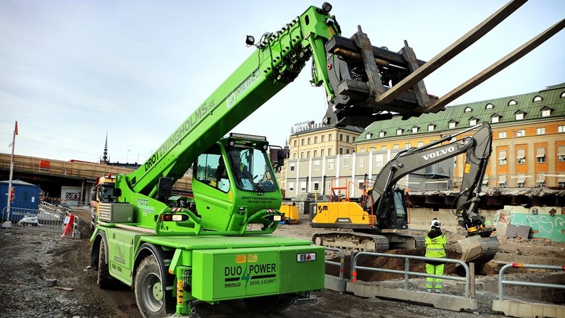Grön teleskopisk gaffeltruck vid stadsbyggarbetsplats med arbetare och grävmaskin.