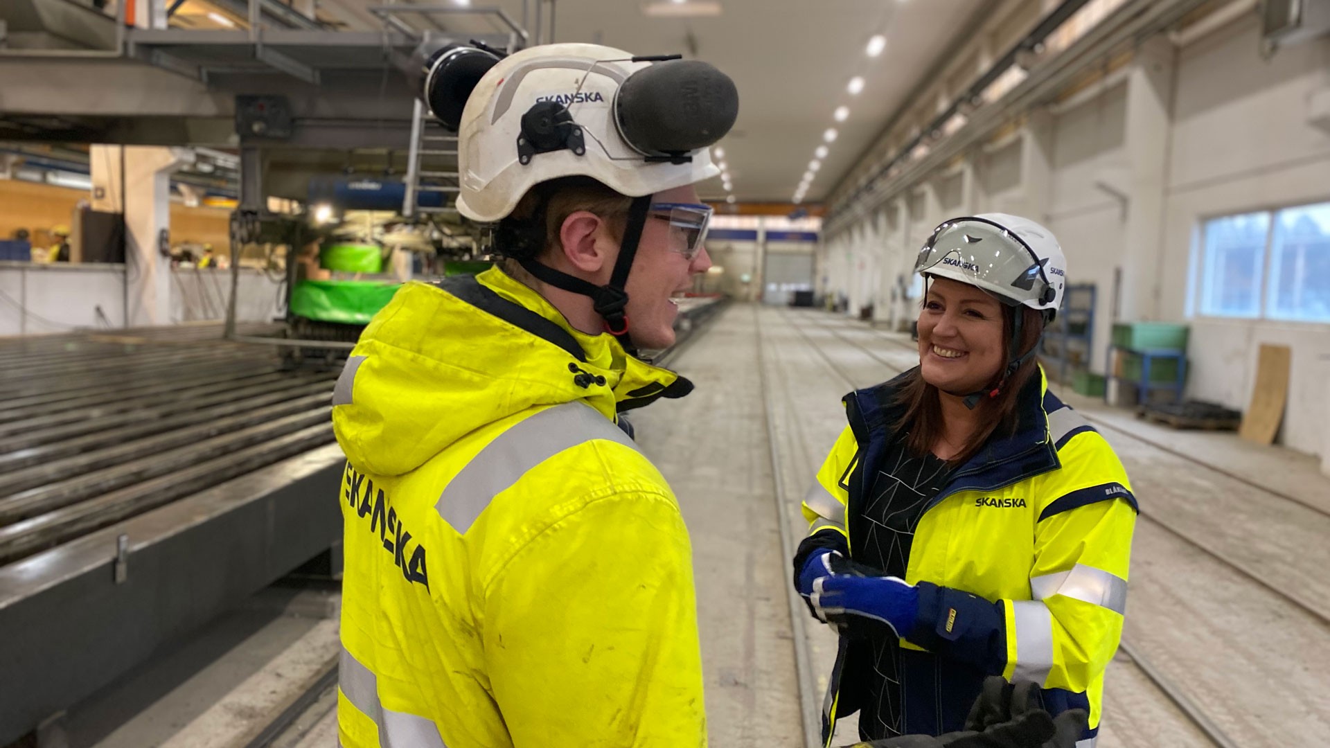 Man i varselkläder i profil som pratar med kvinna i varselkläder. De står på fabriksgolvet med liggande pålar bakom sig.