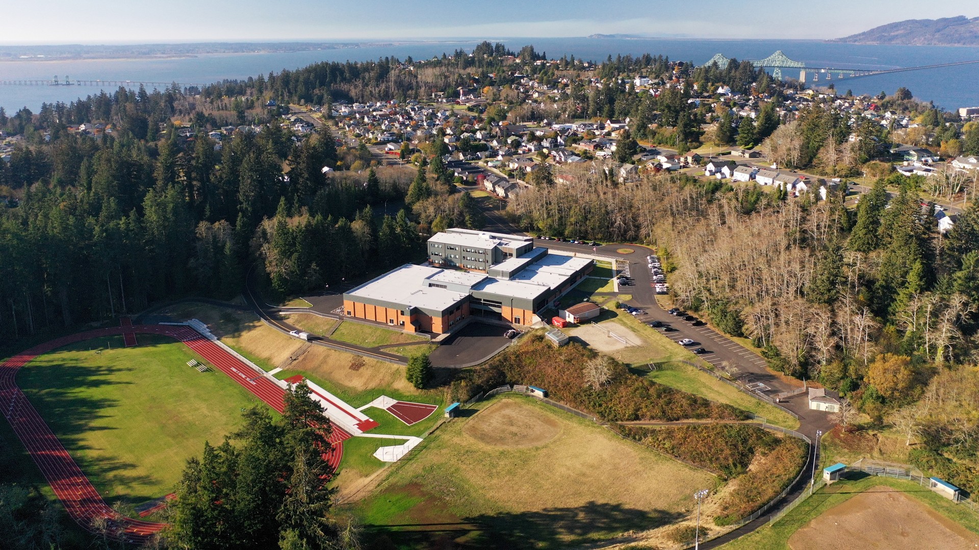 Aerial view of a school surrounded by lush greenery and a track field in a coastal town setting.