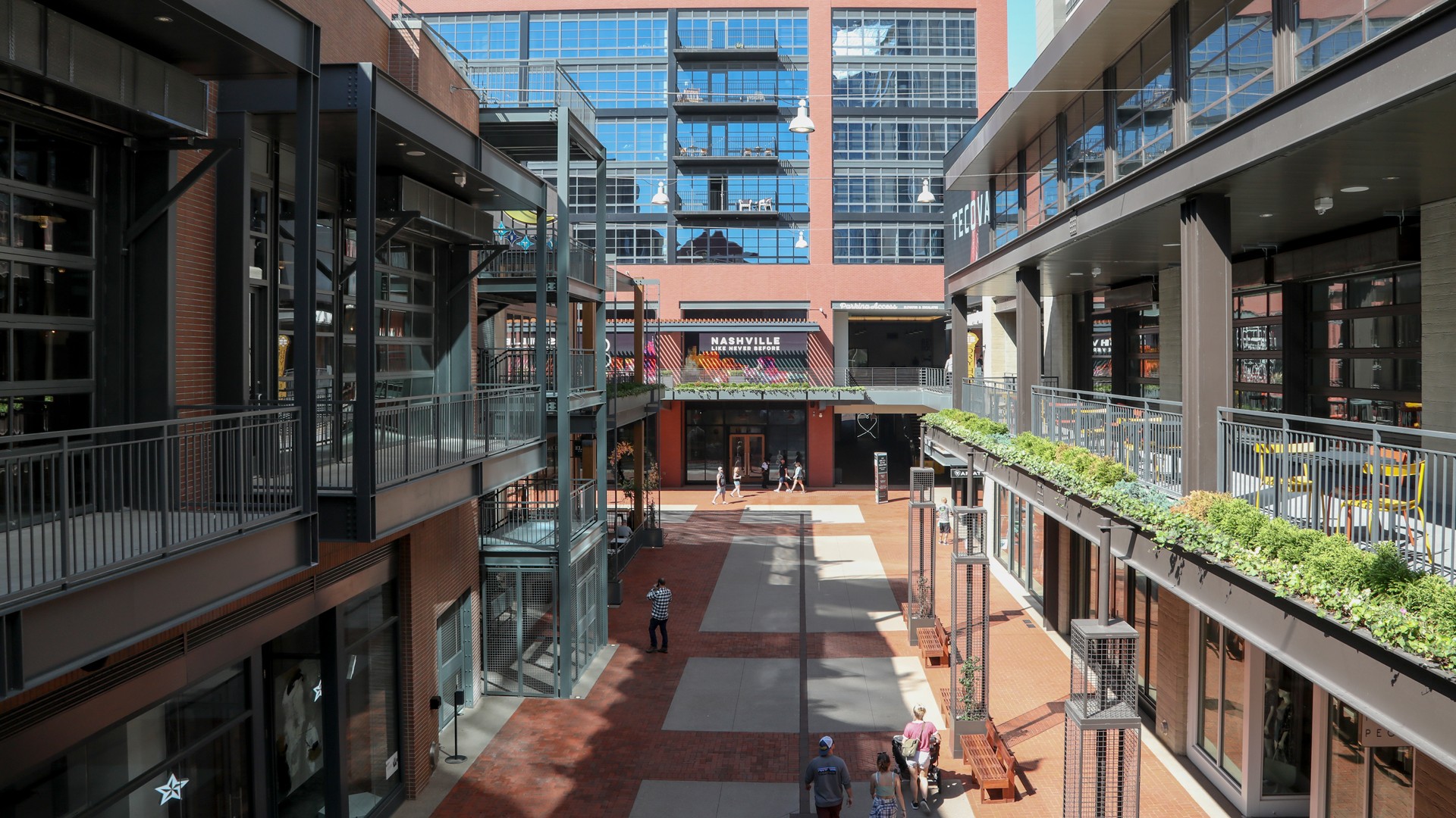 Modern Urban Shopping Center with Walkways and Balconies Modern urban shopping area with balconies, plants, and people walking through a paved pathway.
