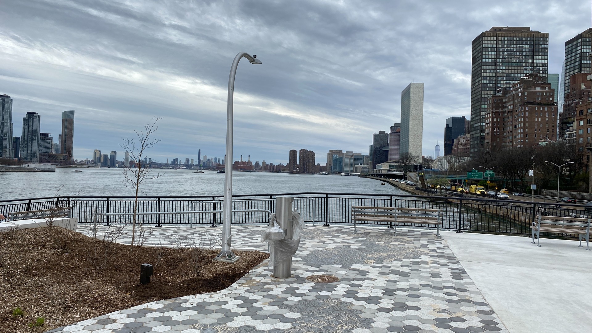 Riverside park with hexagonal tiles, benches, and city skyline under cloudy sky.