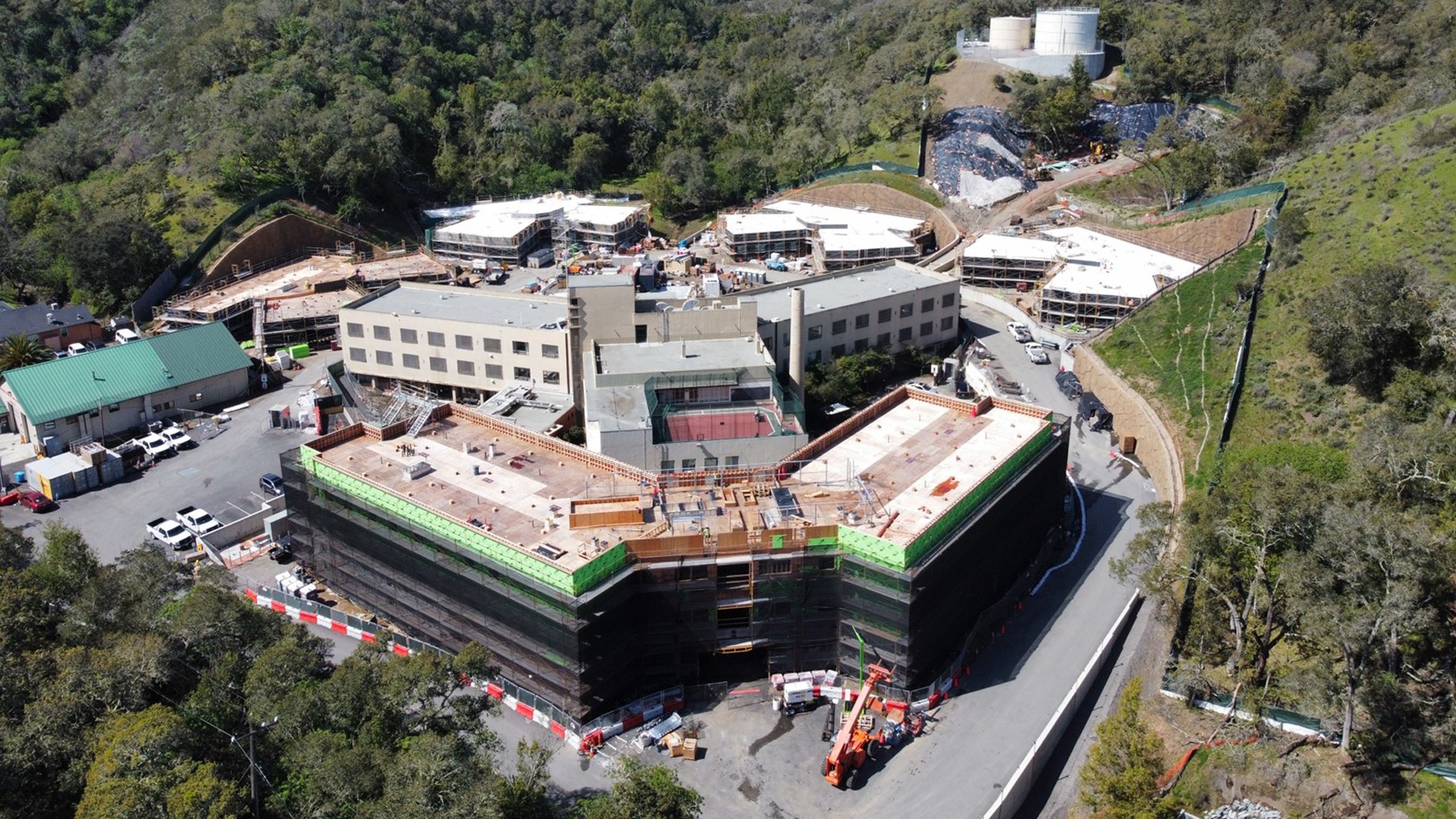 Modern Hospital Construction Amidst Scenic Greenery Aerial view of a large construction site surrounded by trees and hills, showcasing multiple buildings.
