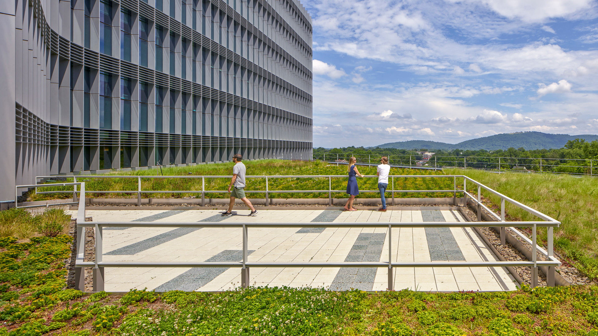 Modern Green Rooftop with Scenic Mountain Views People walking on a rooftop garden with a modern building and scenic mountain backdrop.