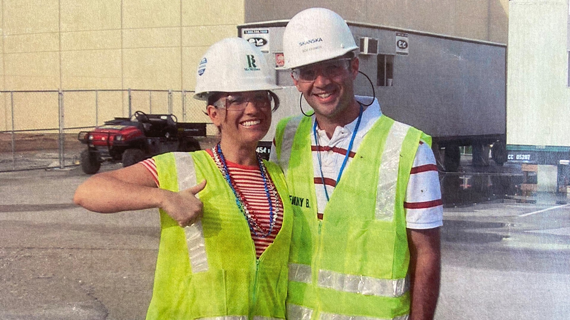 Two construction workers wearing safety vests and helmets posing outdoors on site.