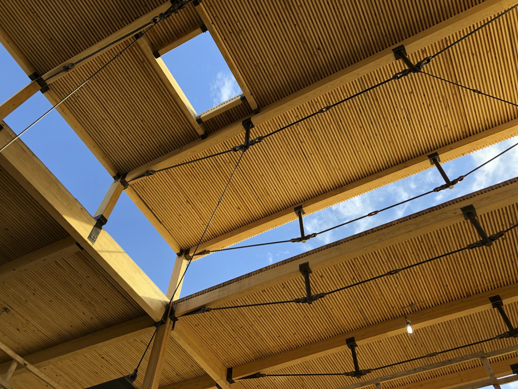 Wooden ceiling structure with beams and cables against a blue sky with clouds.