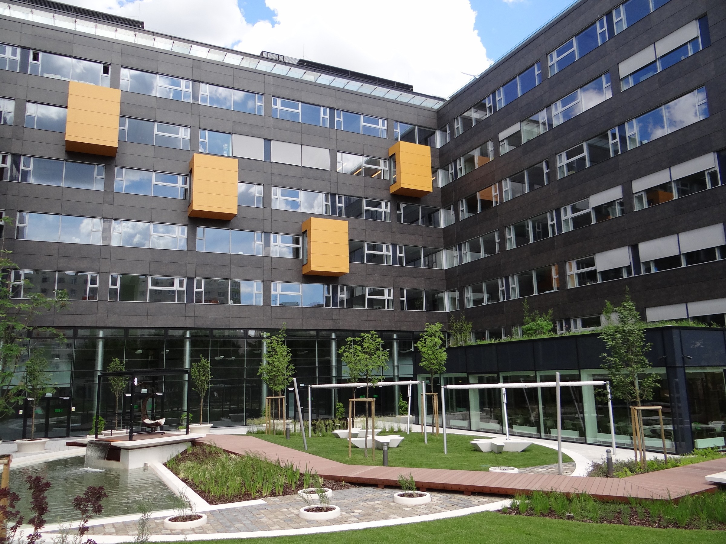 Modern irodaépület egyedi sárga kiemelkedésekkel és zöld növényzettel kialakított udvarral.