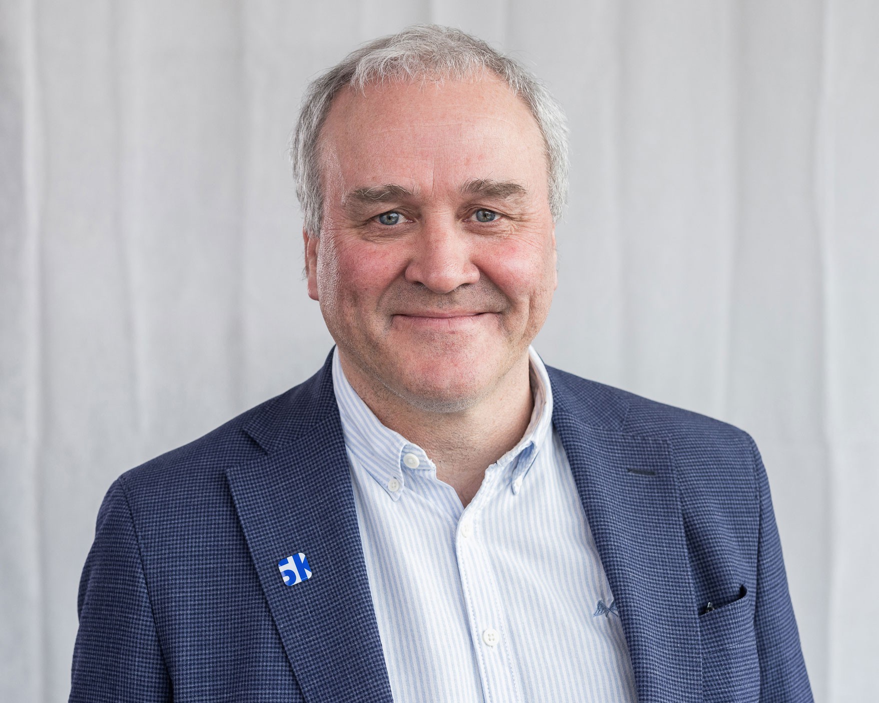 Mann i en blå rutete blazer og hvit skjorte mot en ensfarget bakgrunn.