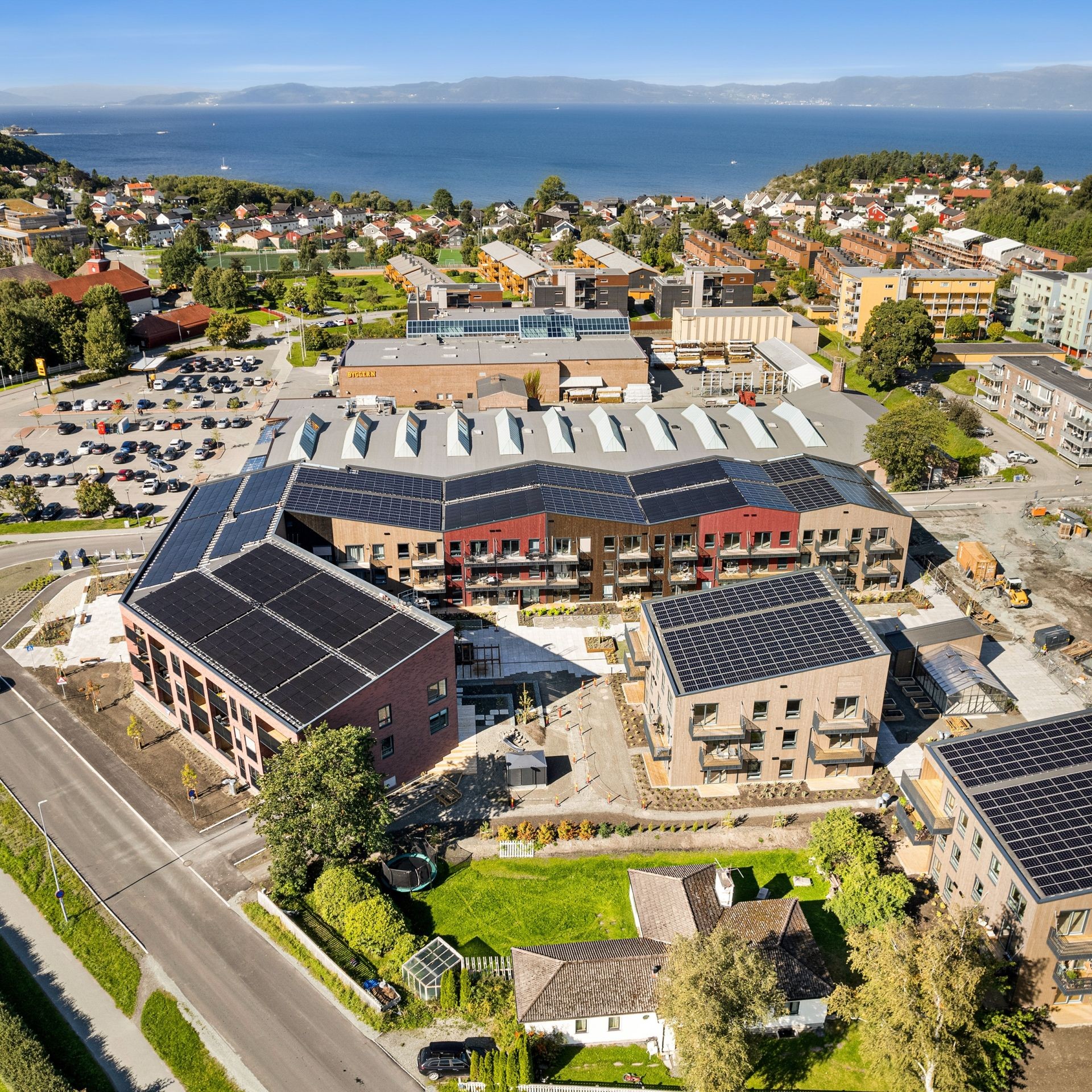 Flyfoto av et moderne leilighetskompleks med solcellepaneler, omgitt av grøntområder og urbant landskap.
