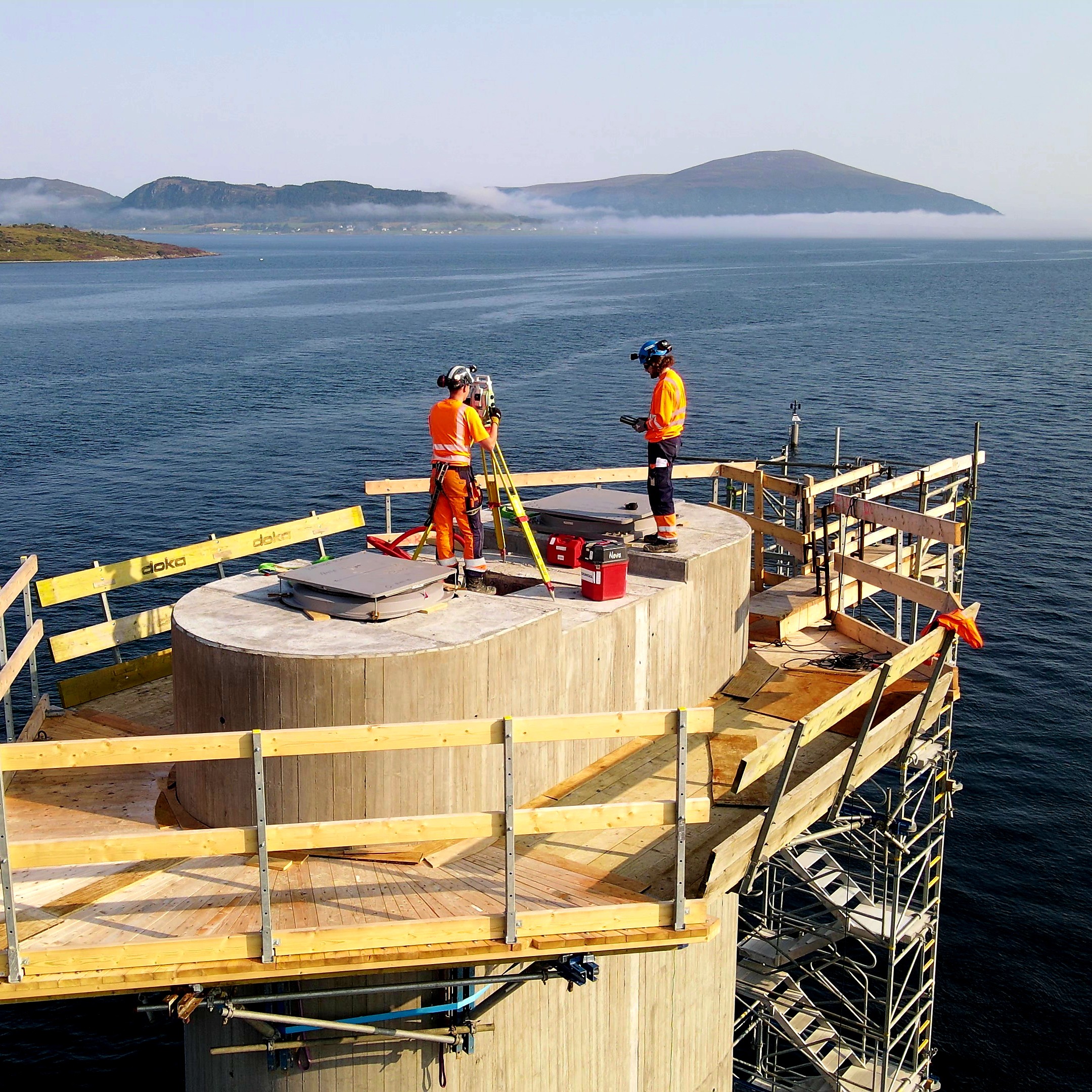 Two construction workers in safety gear using surveying equipment on a coastal platform.