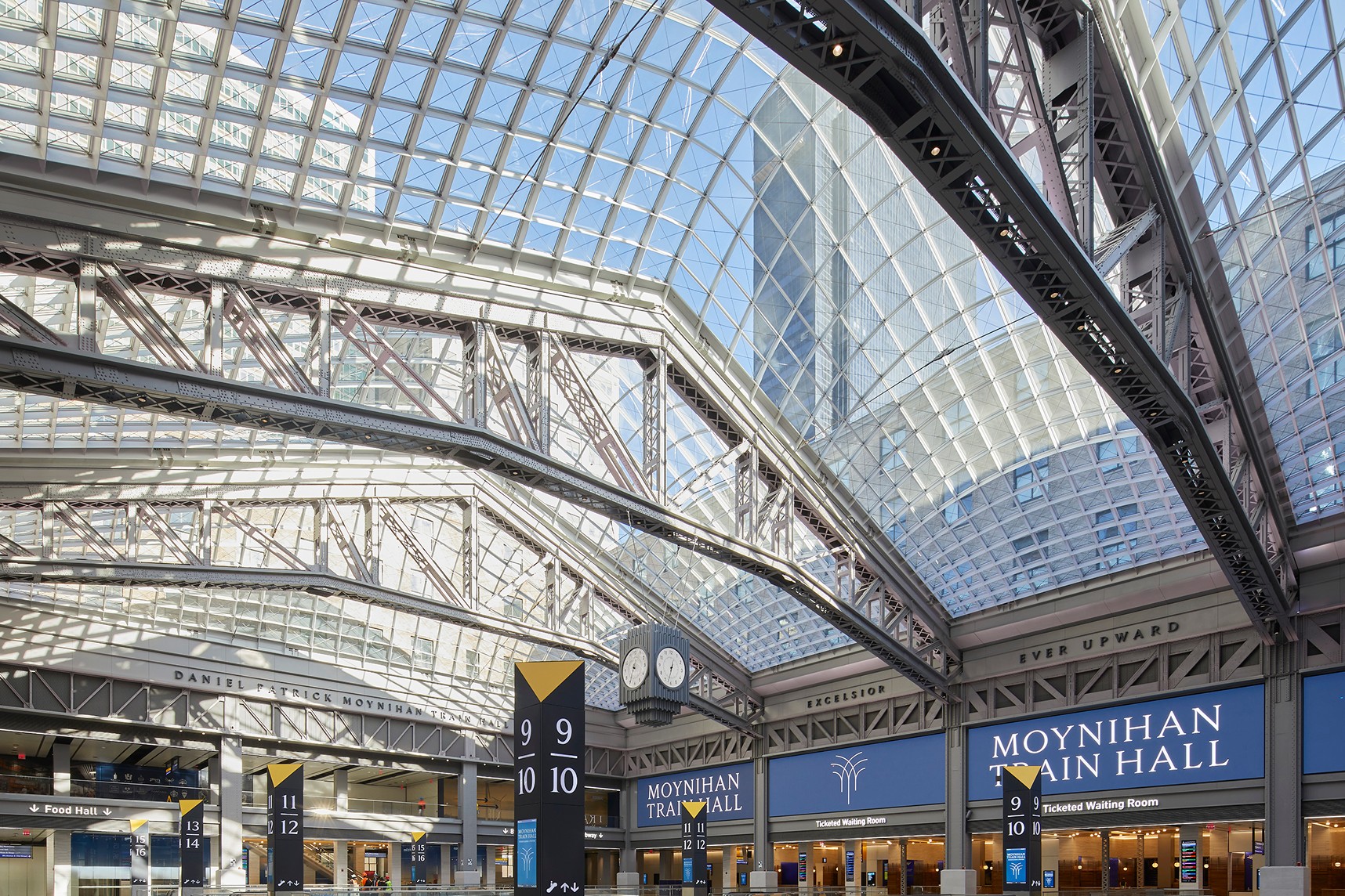 Interior view of Moynihan Train Hall with glass ceiling, signage, and train platforms visible.