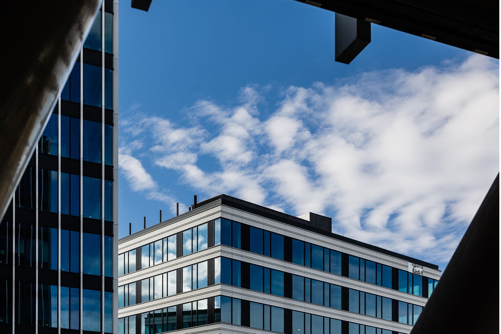 Modern office buildings against a blue sky with scattered clouds, framed by dark architectural elements.