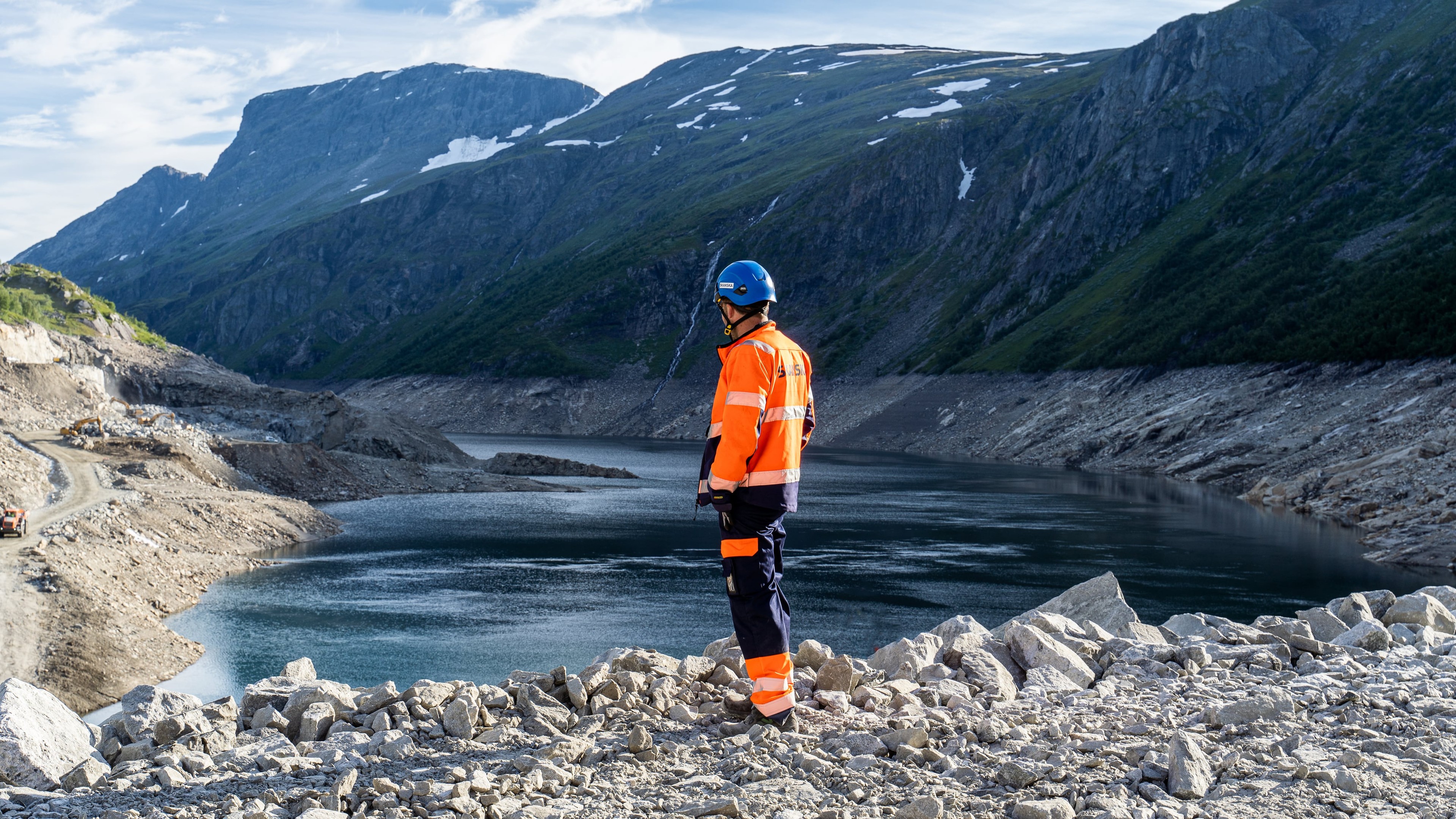 Worker in safety gear overlooking rocky terrain and water in mountainous landscape.