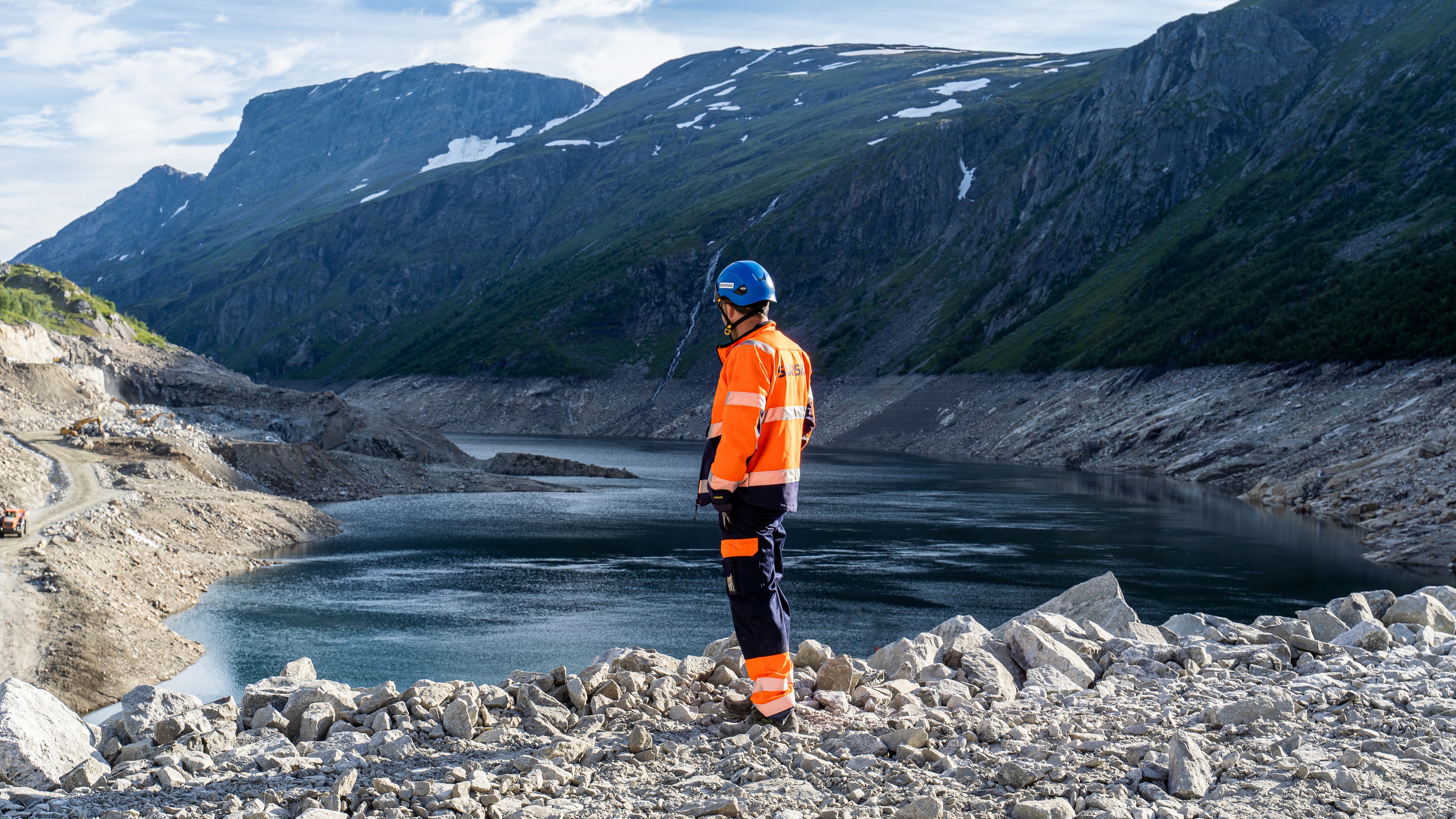 Worker in safety gear overlooking rocky terrain and water in mountainous landscape.