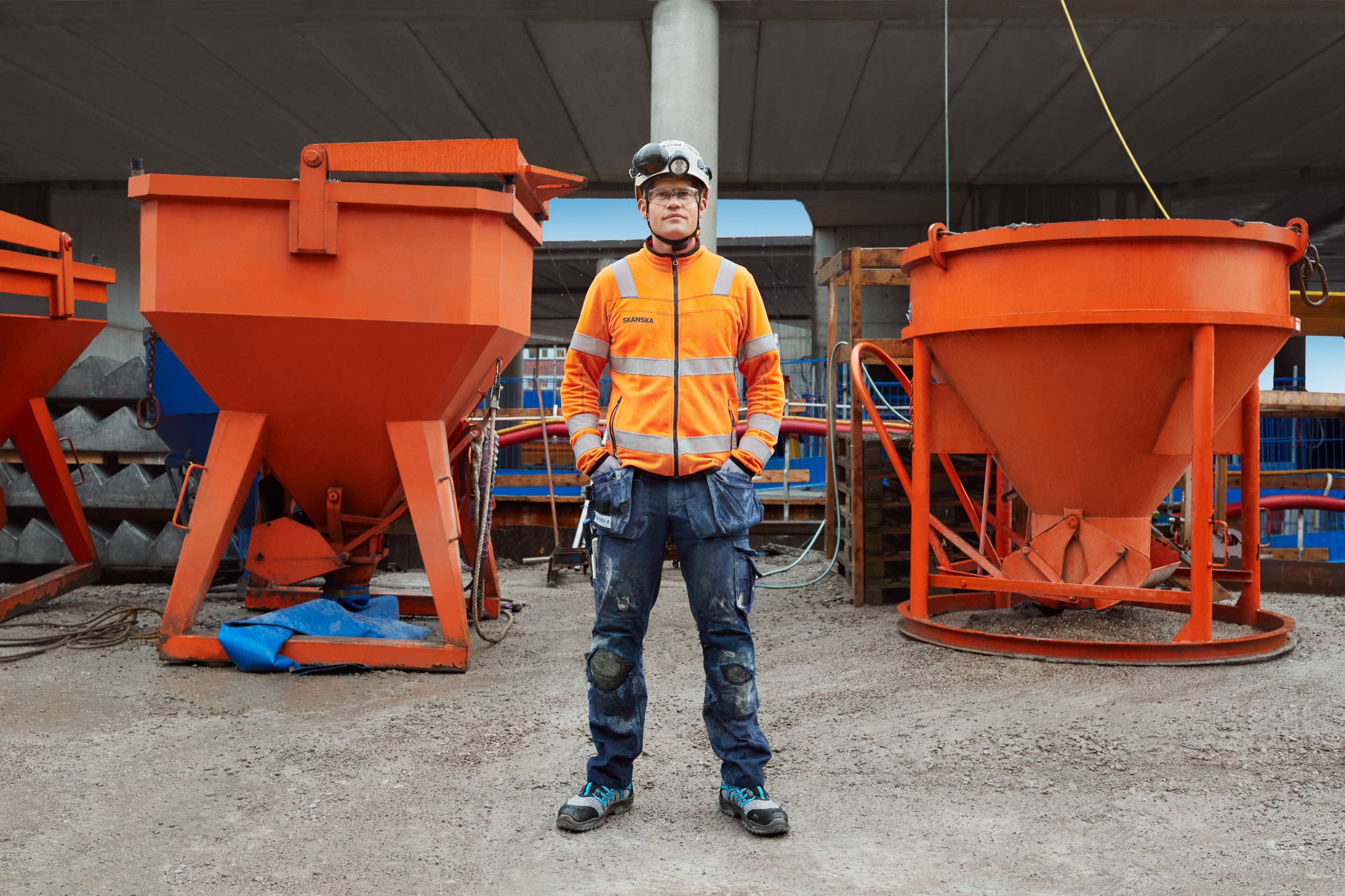 Construction worker in safety gear standing between large orange concrete mixers on site.