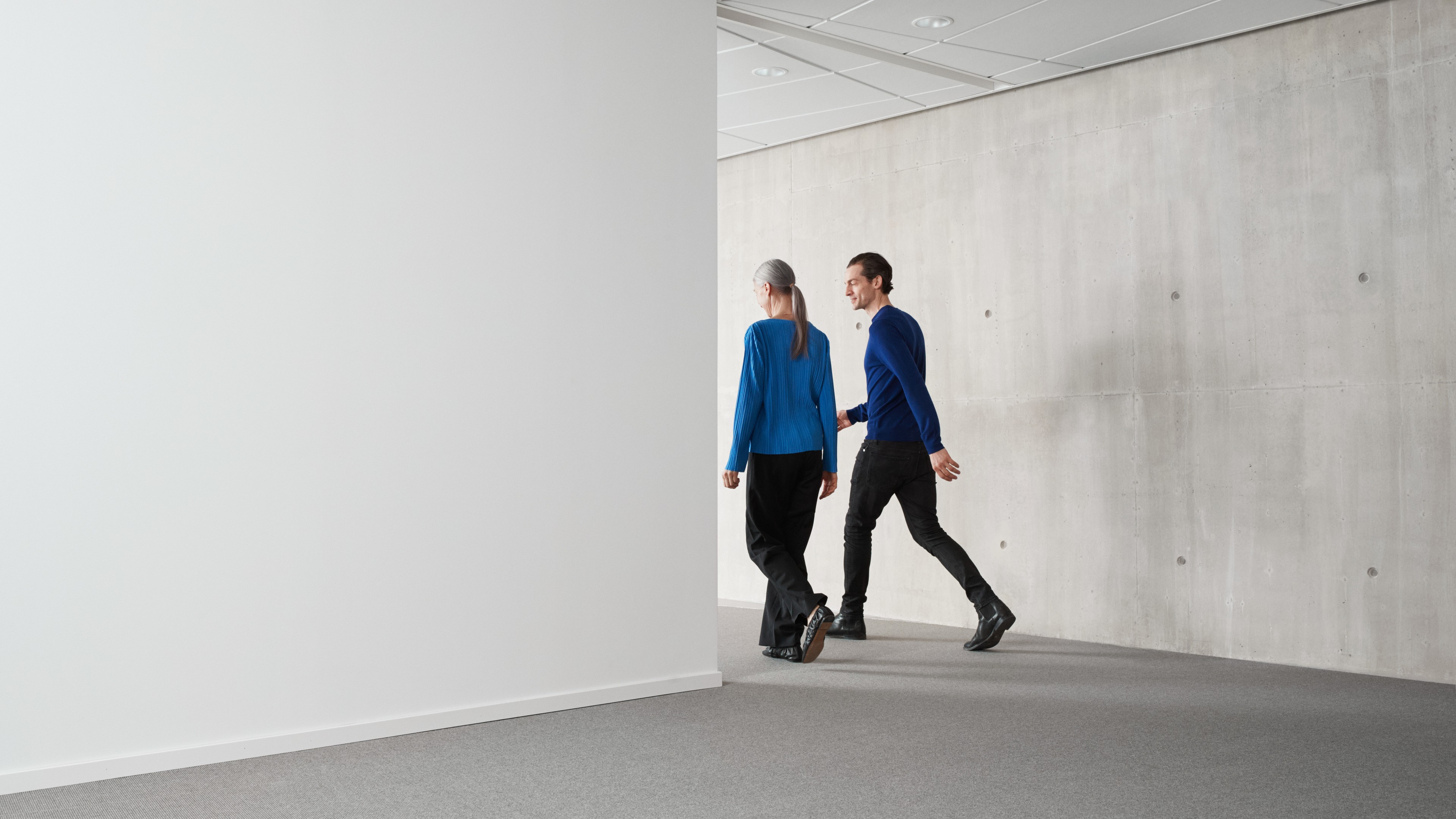 Two people in blue sweaters walking in a modern minimalist office hallway with gray walls.