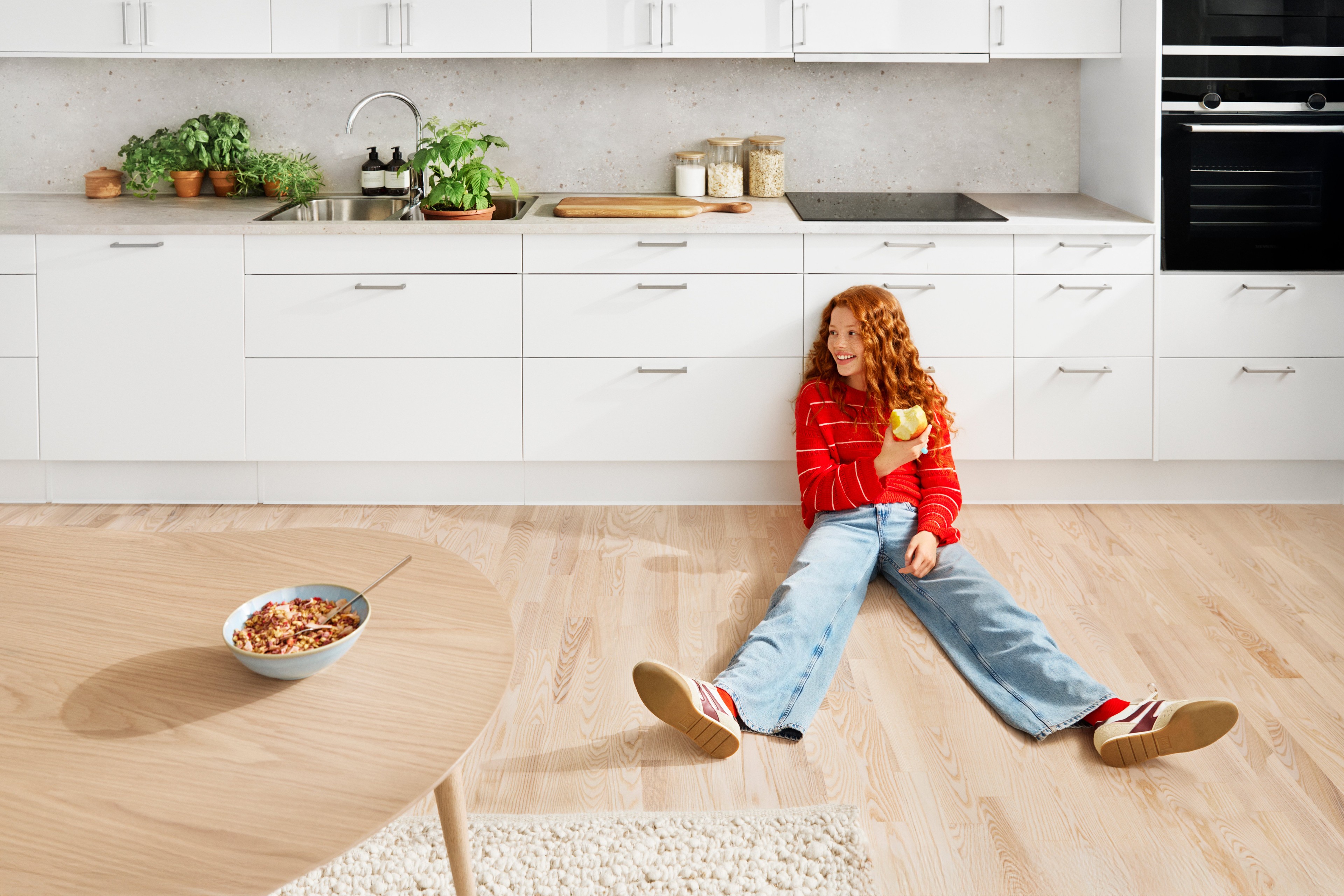 Child in red sweater eating apple while sitting on kitchen floor near white cabinets.