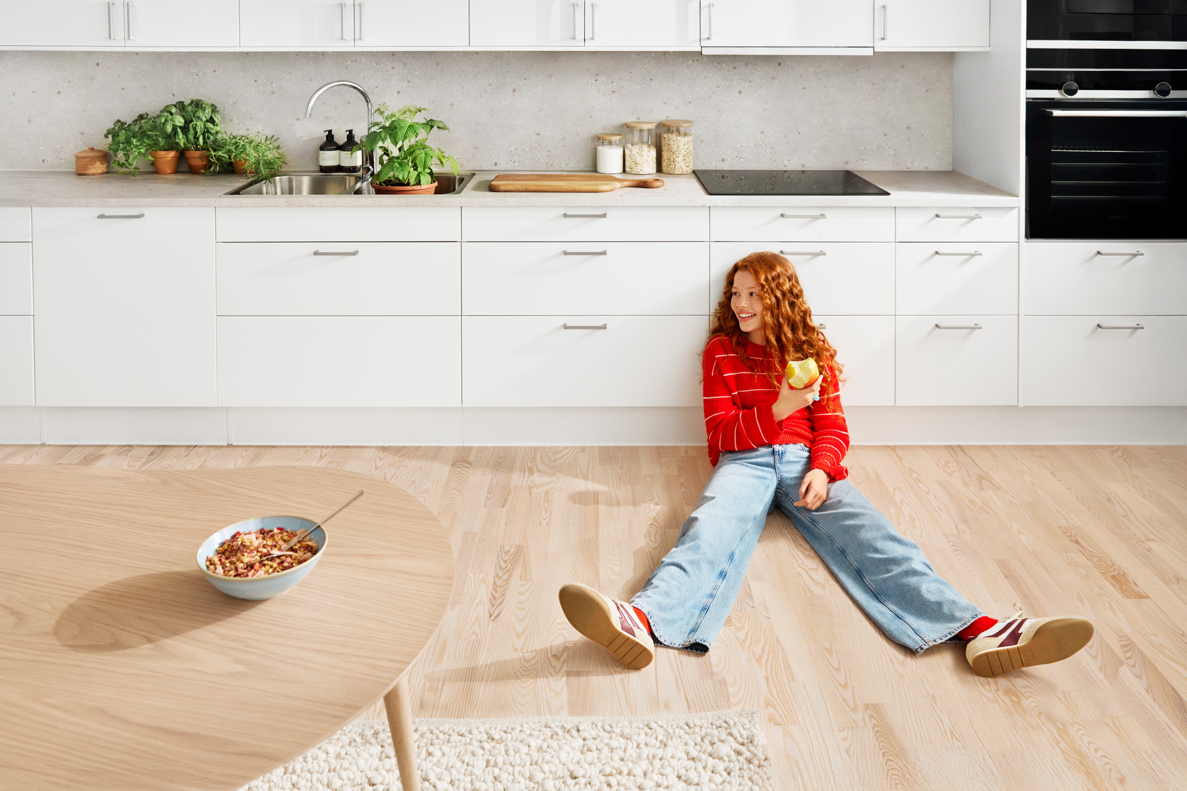 Child in red sweater eating apple while sitting on kitchen floor near white cabinets.