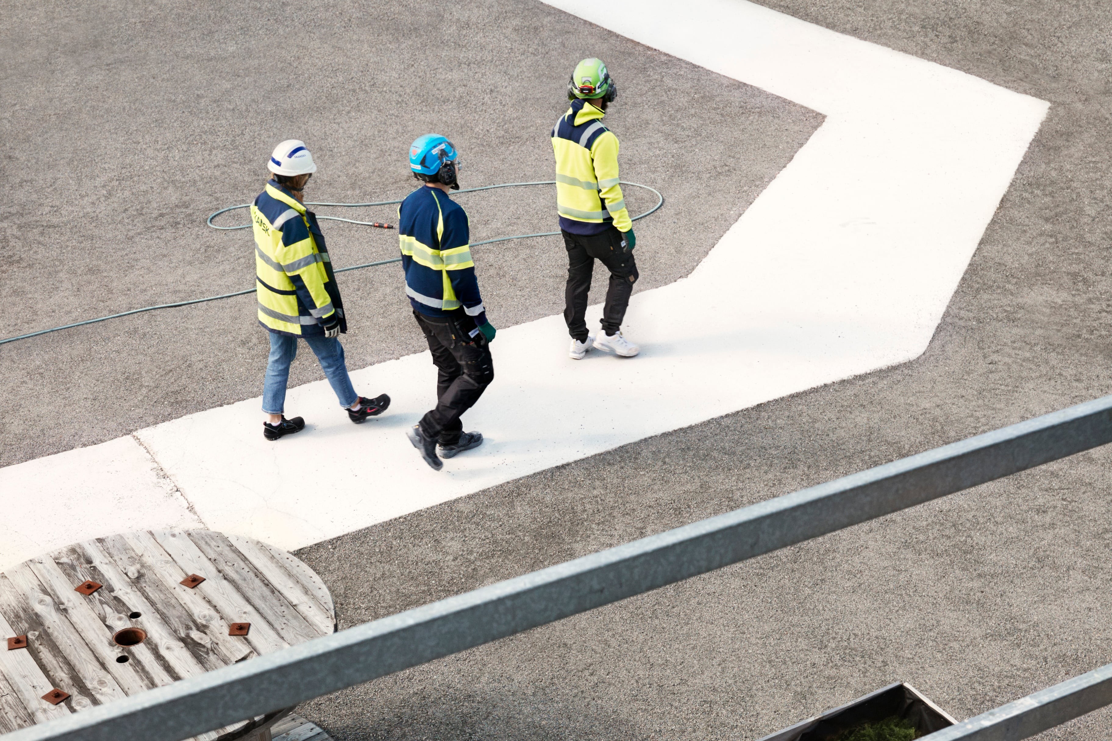 Three construction workers in safety gear walk on a marked path at a worksite.