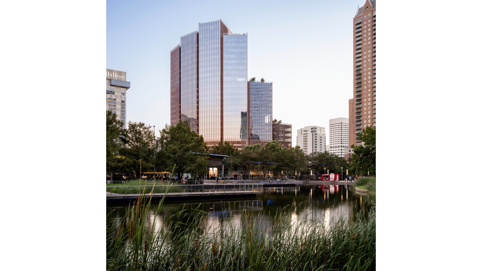 Modern skyscrapers and lush greenery reflected in a tranquil pond at sunset in an urban park.