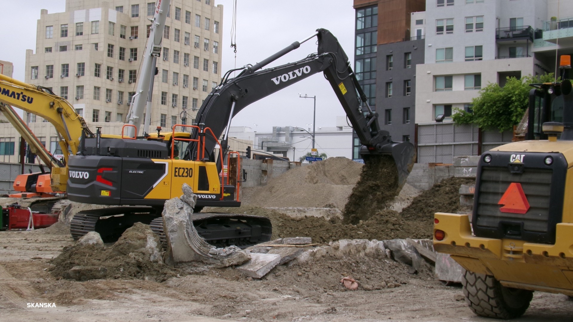 Excavators from Volvo and Komatsu operate at a construction site surrounded by buildings.