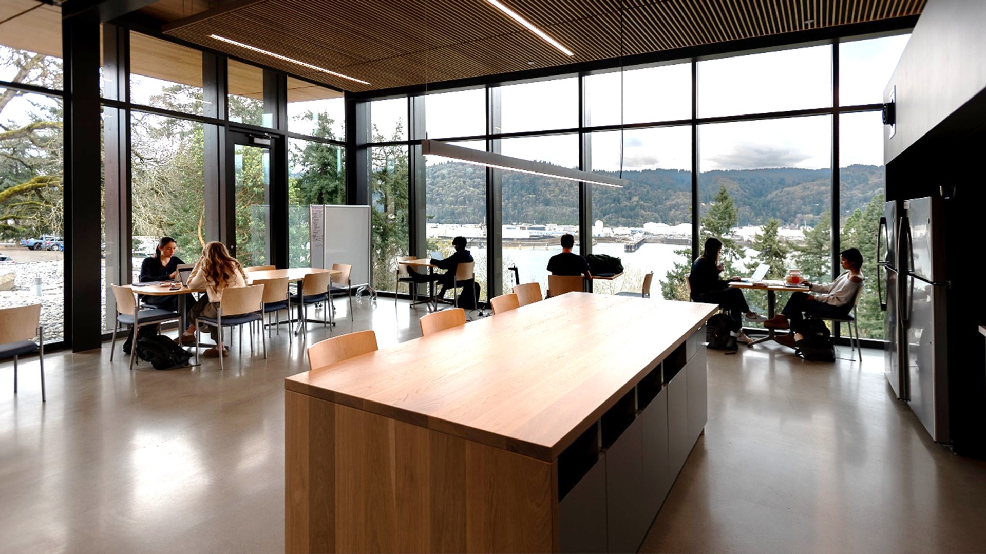 The University of Portland – Shiley-Marcos Center for Design & Innovation Modern office break room with large windows, communal tables, and people enjoying the view.