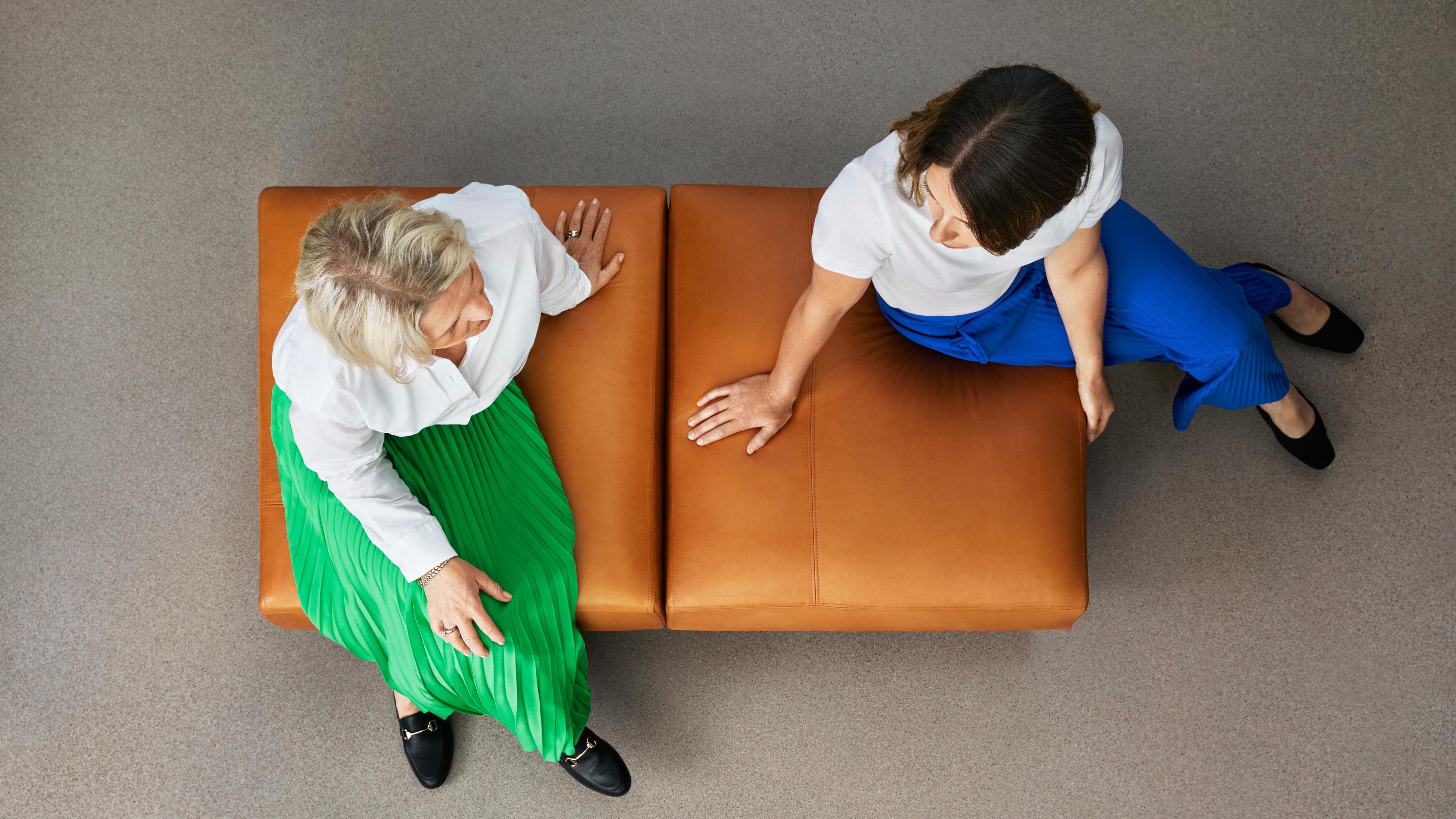 Two women seated on a brown bench, one in a green skirt and the other in blue pants, viewed from above.