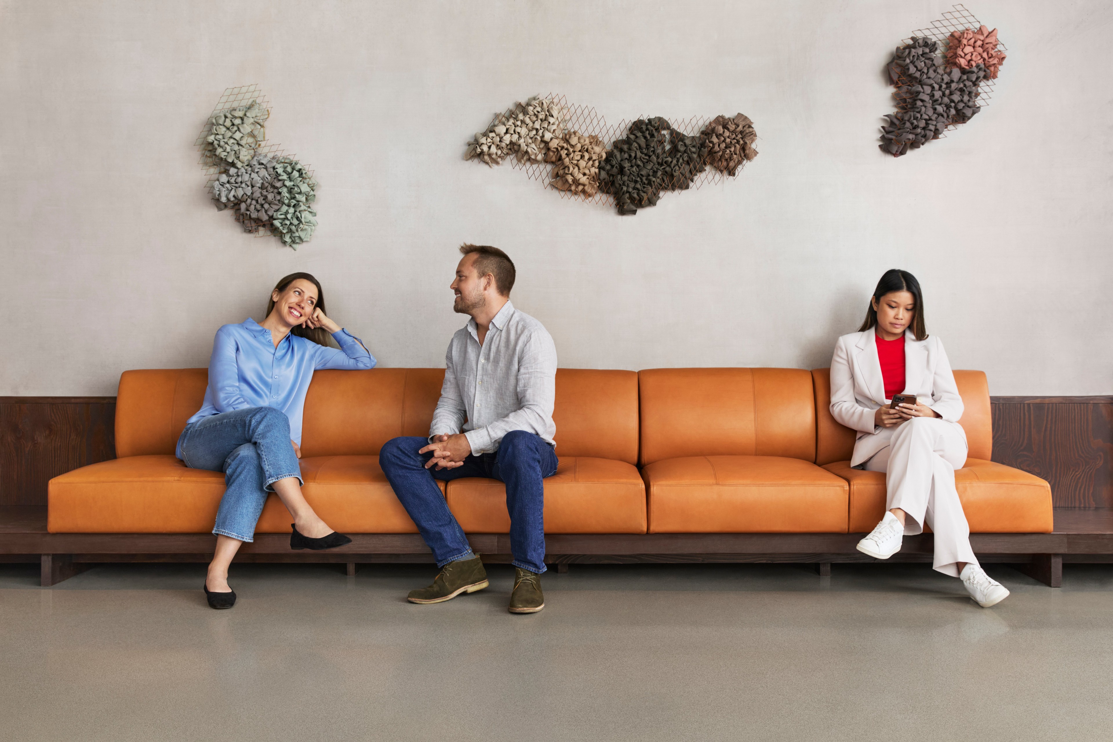 Three people sitting on a modern orange couch in a stylish waiting room with wall art.