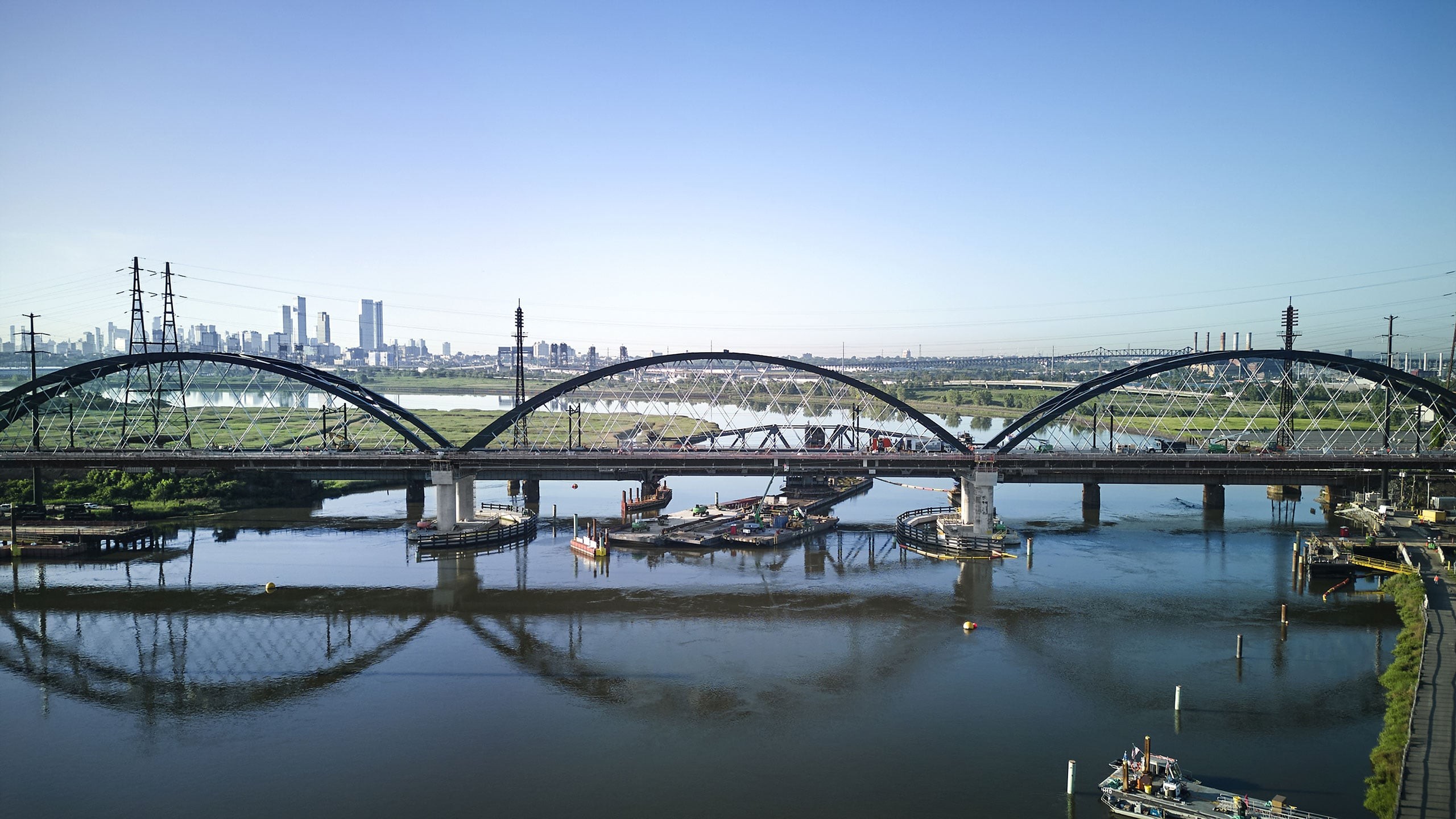 Steel arch bridge over calm river with city skyline and clear sky in the background.