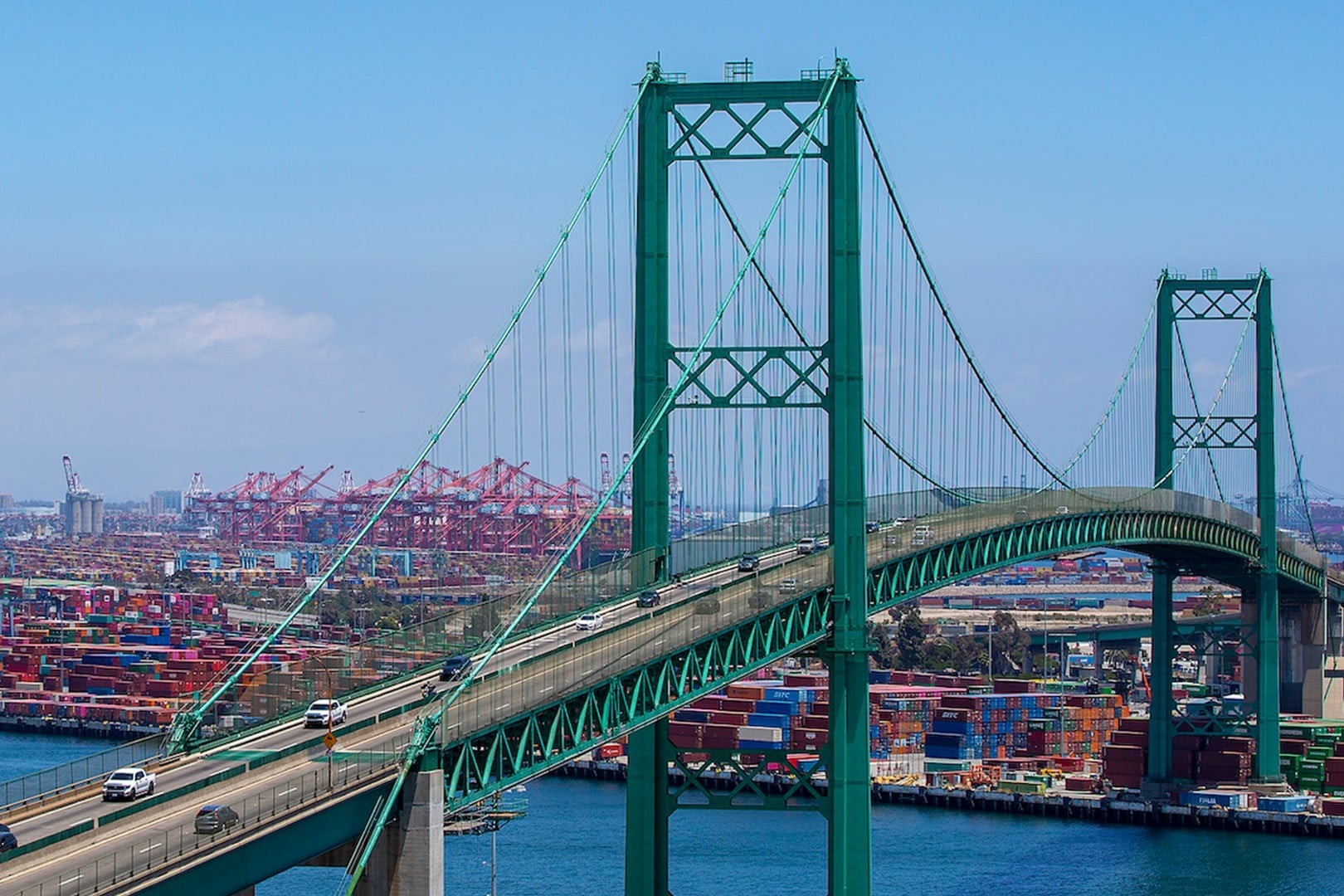 Suspension bridge over water with shipping containers and industrial cranes in background.