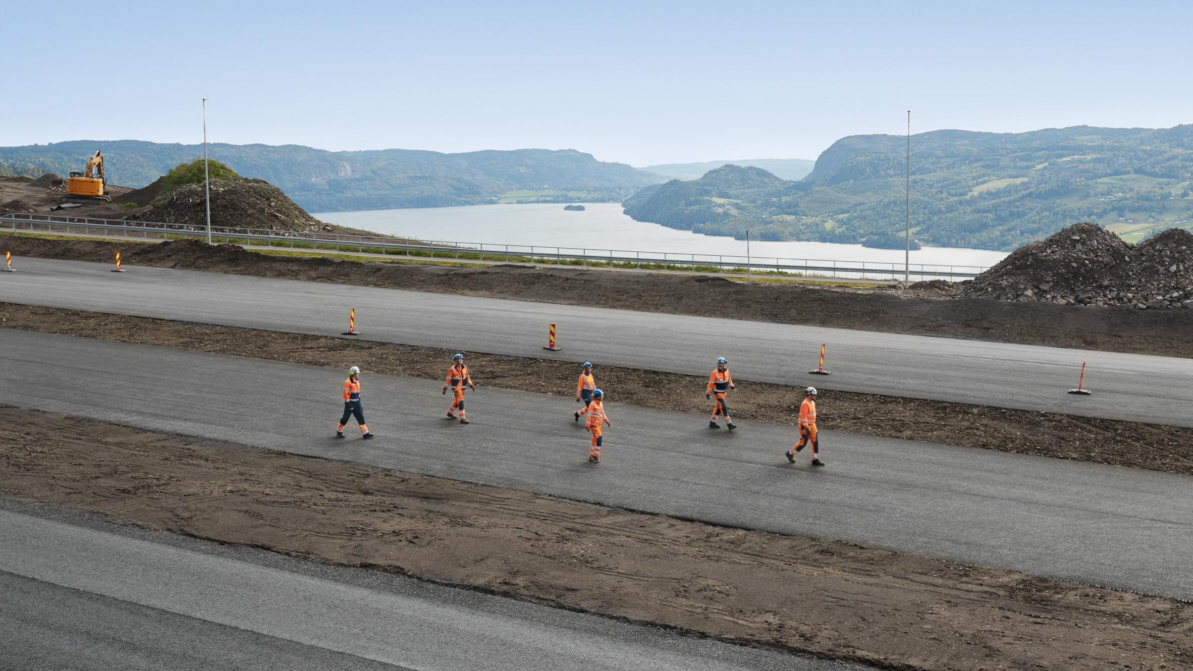 Construction workers in safety gear walk on a newly paved road with hills and water in background.