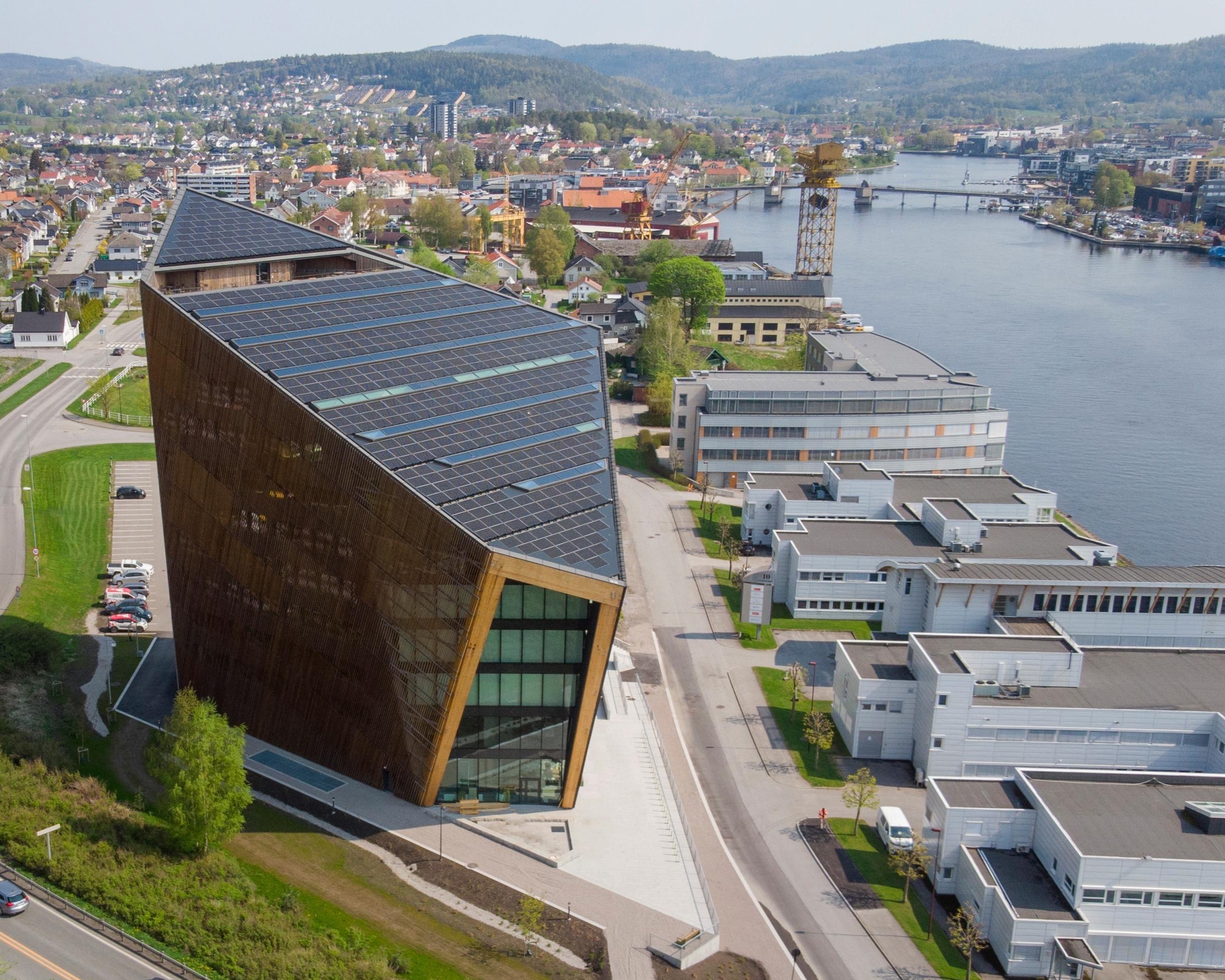 Modern triangular building with solar panels by a riverside in a scenic townscape setting.