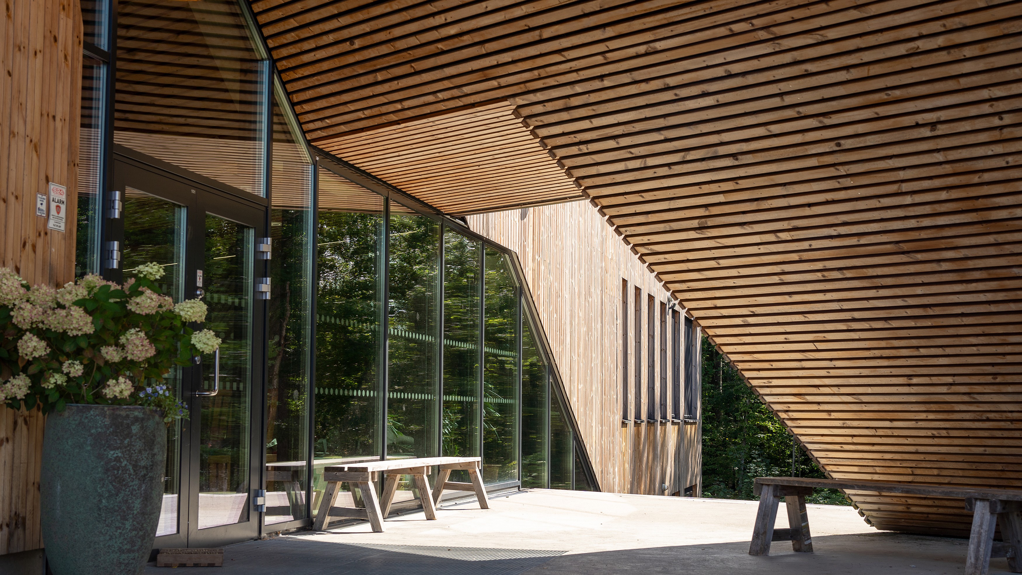 Modern wooden building with angled roof, glass walls, and benches in a sunlit outdoor space.