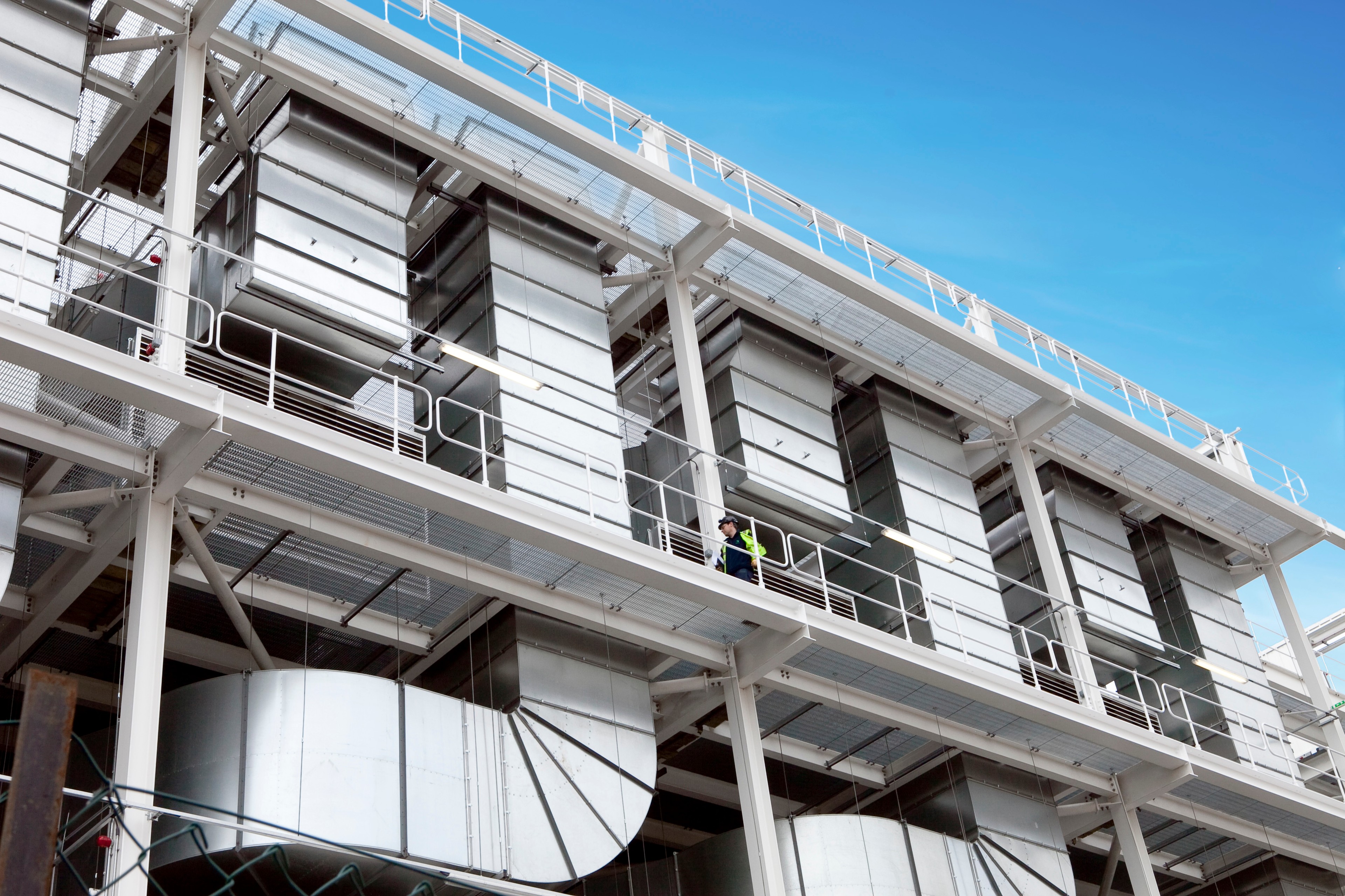Industriellt HVAC-system med stora metallkanaler och en arbetare i säkerhetsutrustning, mot en klar blå himmel.