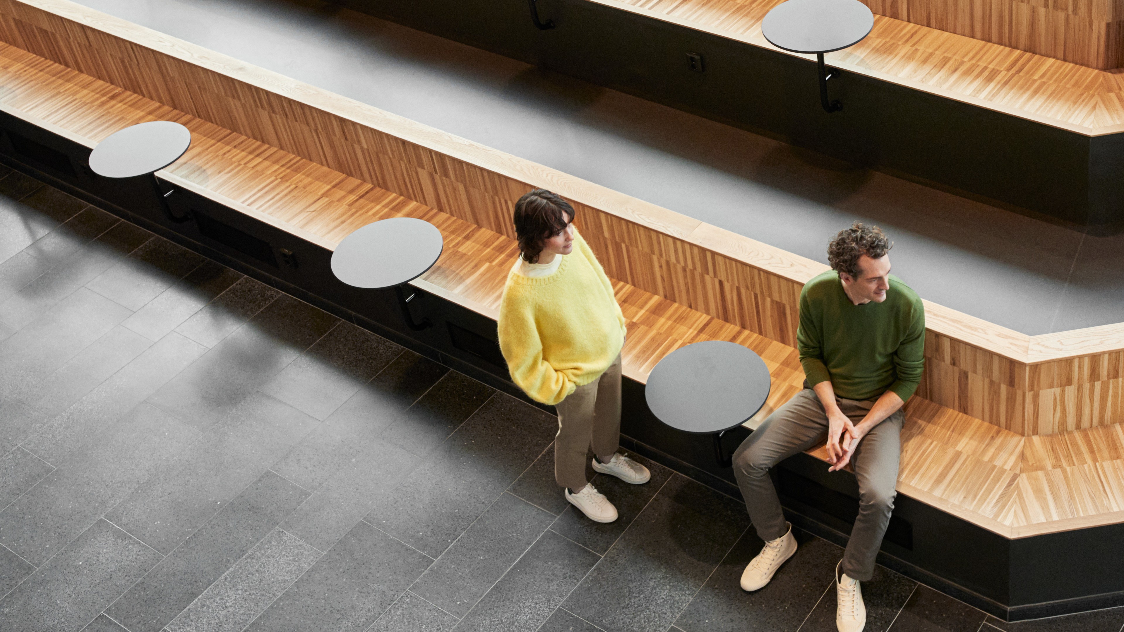 Two people in casual clothing converse on tiered wooden seating with round tables.