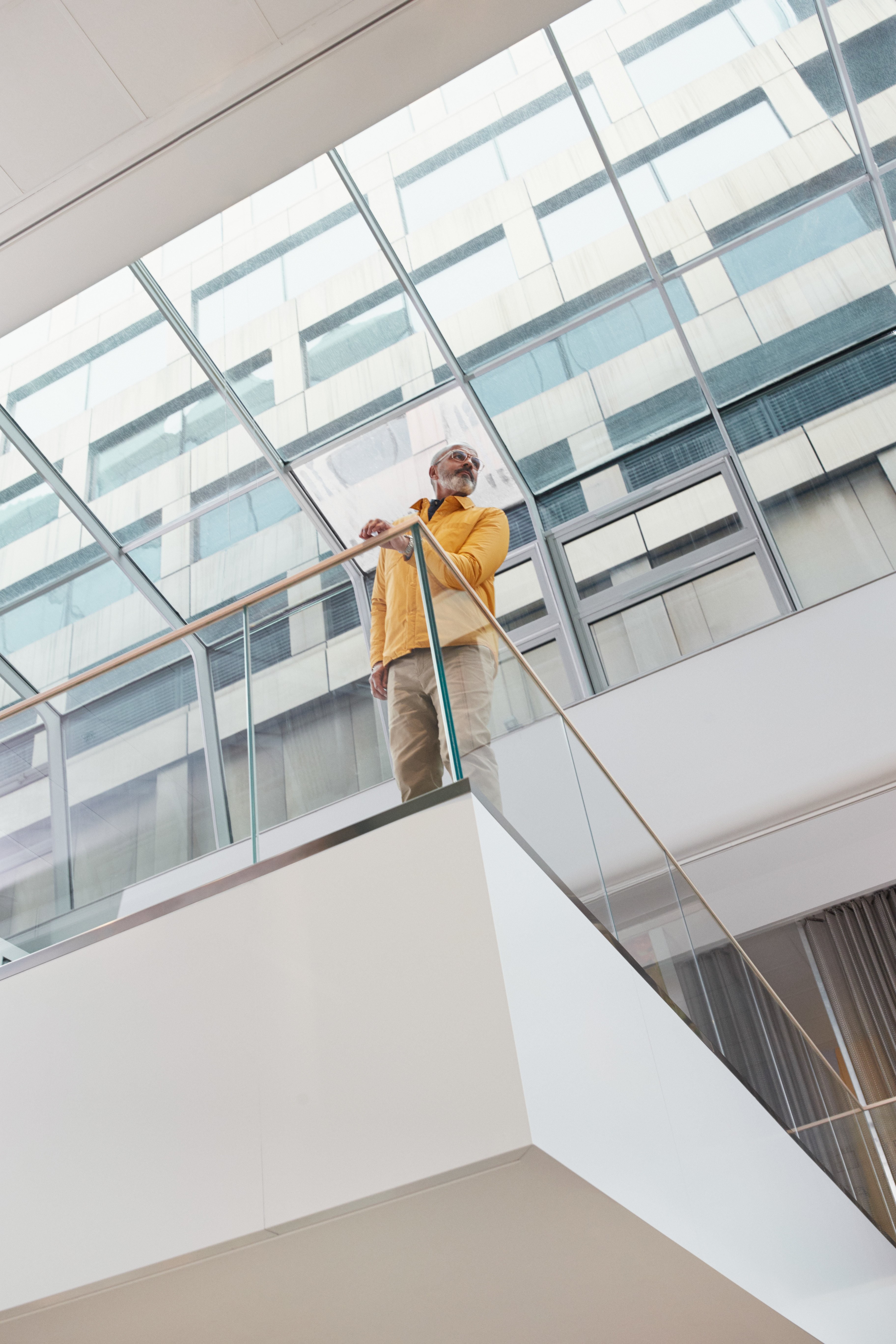 A man in a yellow jacket stands on a modern glass balcony, with a large building visible behind.